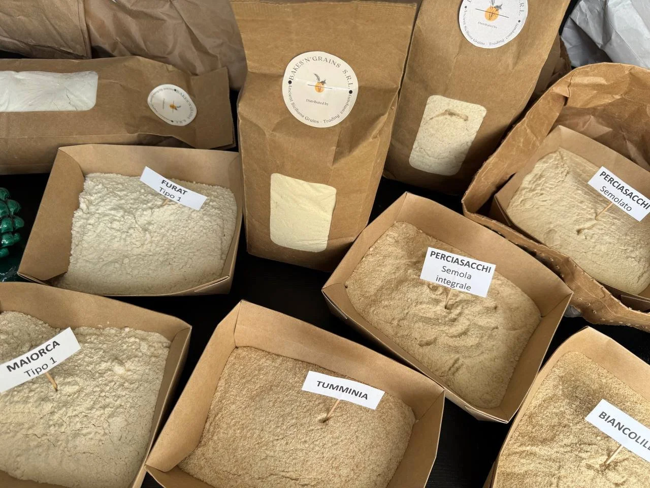 Varie farine in confezioni di carta, con etichette che indicano i tipi di farine come Furat, Perciasacchi, Maiorca, Tumminia, Biancolilla, in un'ambientazione rustica.