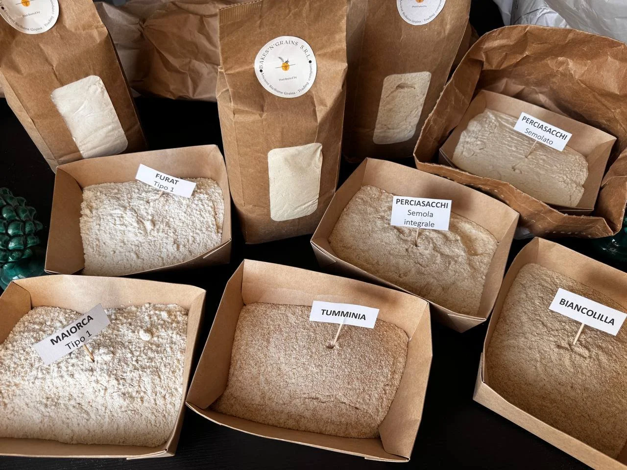 Confezioni di farine e cereali in sacchi di carta con etichette che indicano i diversi tipi, tra cui Furat, Perciasacchi, Maiorca, Tumminia, Bignolilla, e varie tipologie di grano e semola.
