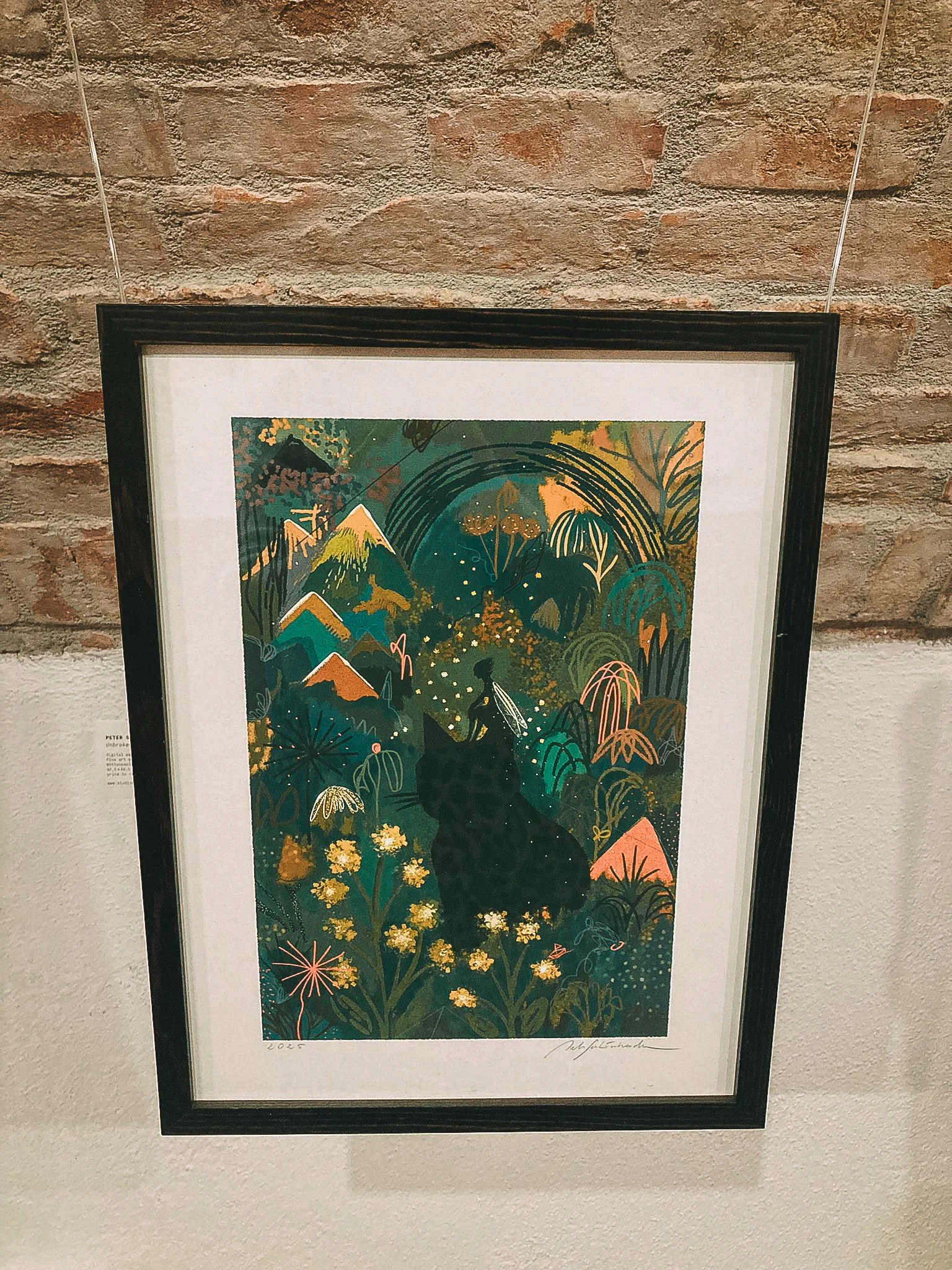 A framed colorful artwork depicting a black cat with a fairy perched on its back, surrounded by lush greenery, mountains, and rainbows.