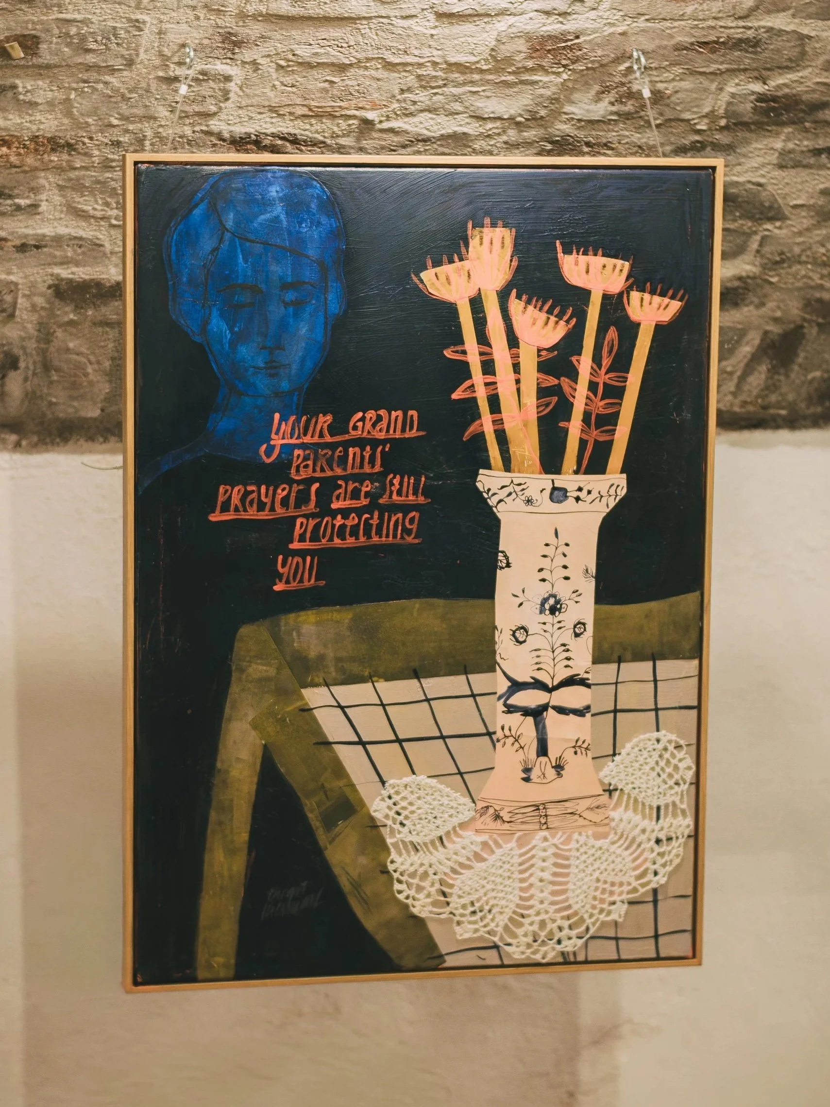 A chalkboard with a blue portrait of a woman, text in red that says 'Your grand 'parents' prayers are still protecting you', a drawing of a vase with pink flowers, and a crocheted doily on a table with a checkered cloth.