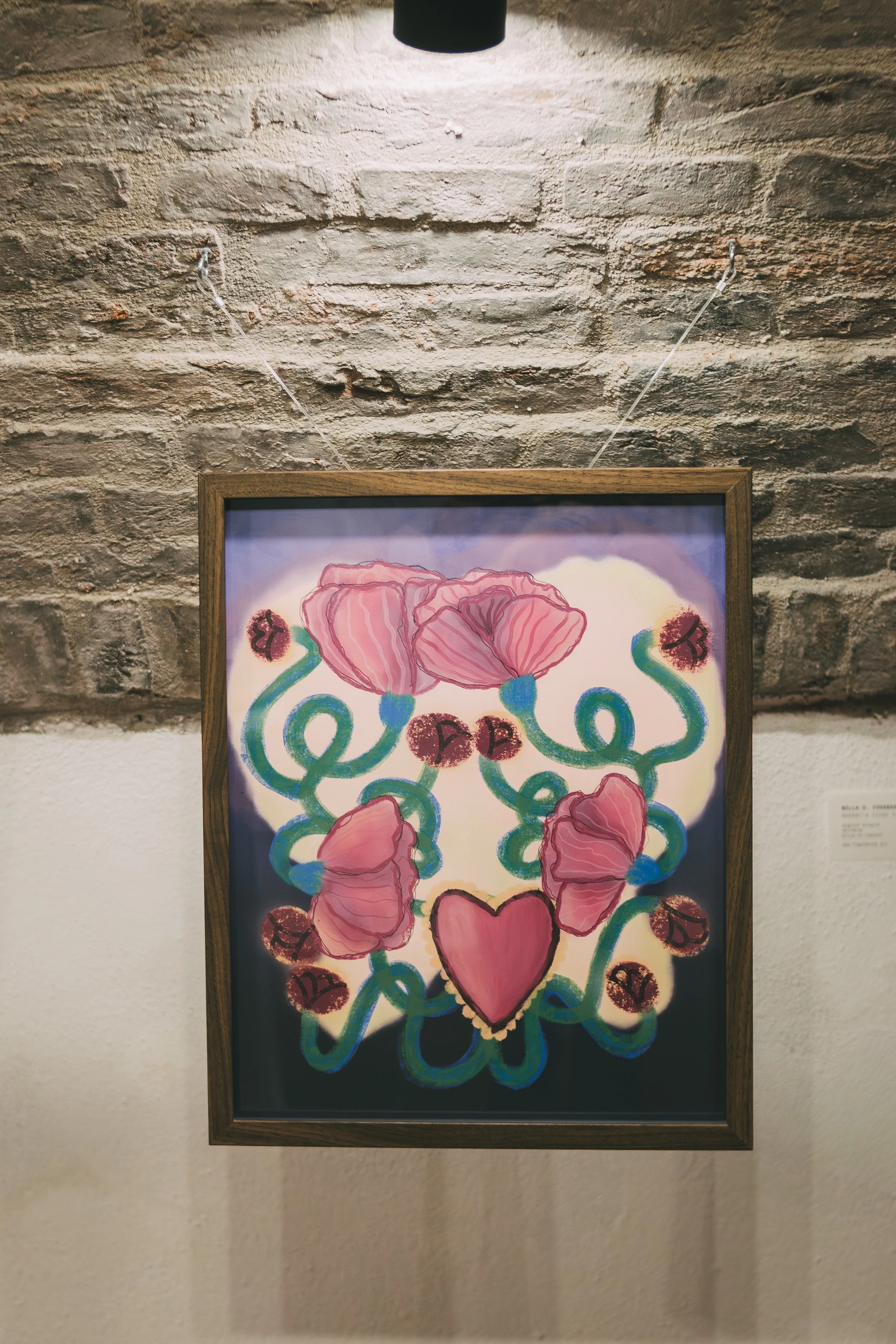 Colorful abstract artwork featuring pink flowers, green swirling stems, and a large pink heart in the center, framed and hung on a brick wall.