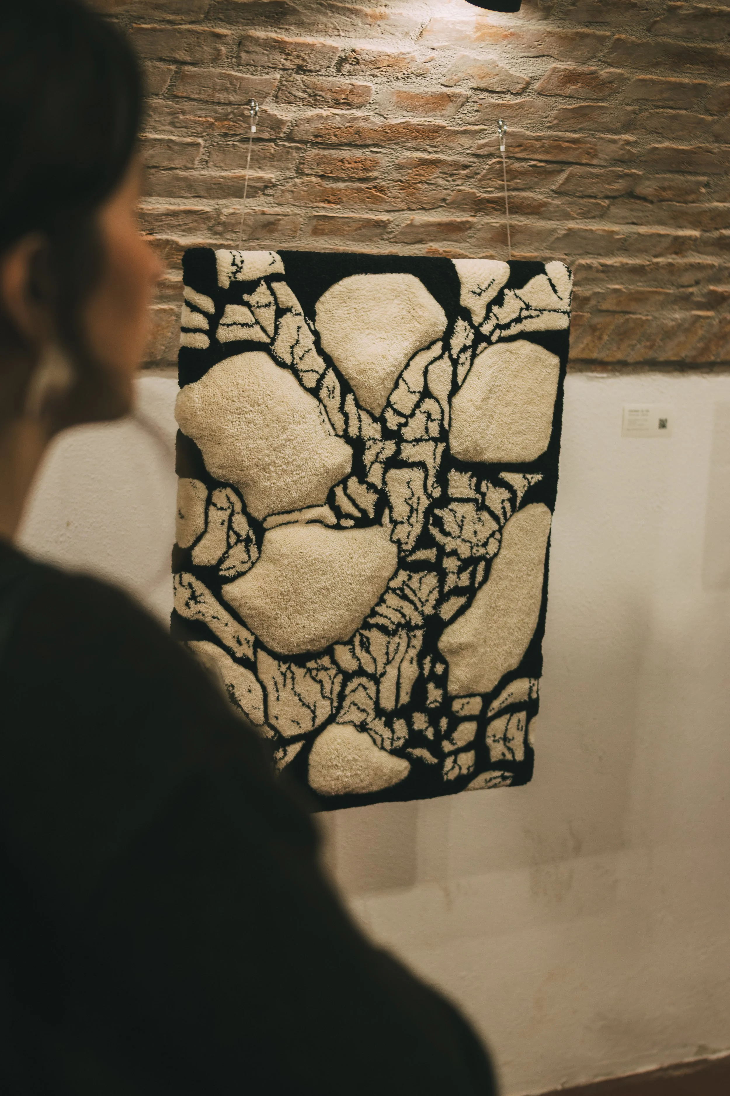 View of a woman standing in front of a textured beige and black abstract textile art piece on a gallery wall with a brick background.