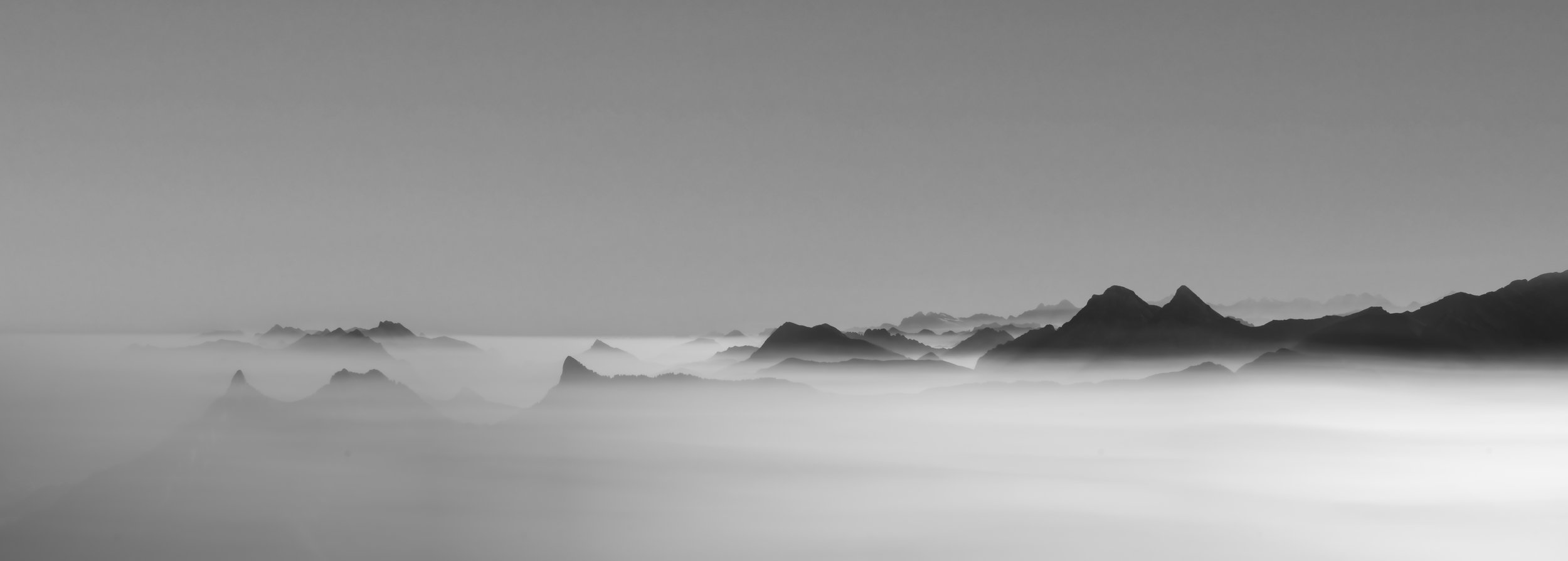 A wide black and white panorama of the Swiss alps peaking above cloud