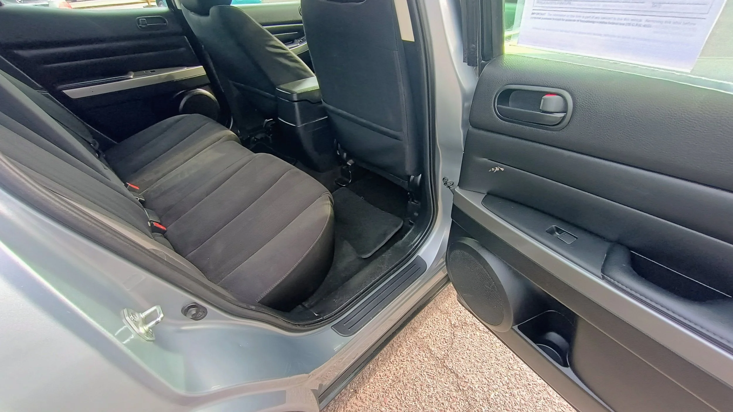 Interior of a vehicle showing the back seat area with black fabric seats, a black floor mat, and the open rear door with black interior paneling and a side window with a paper attached.