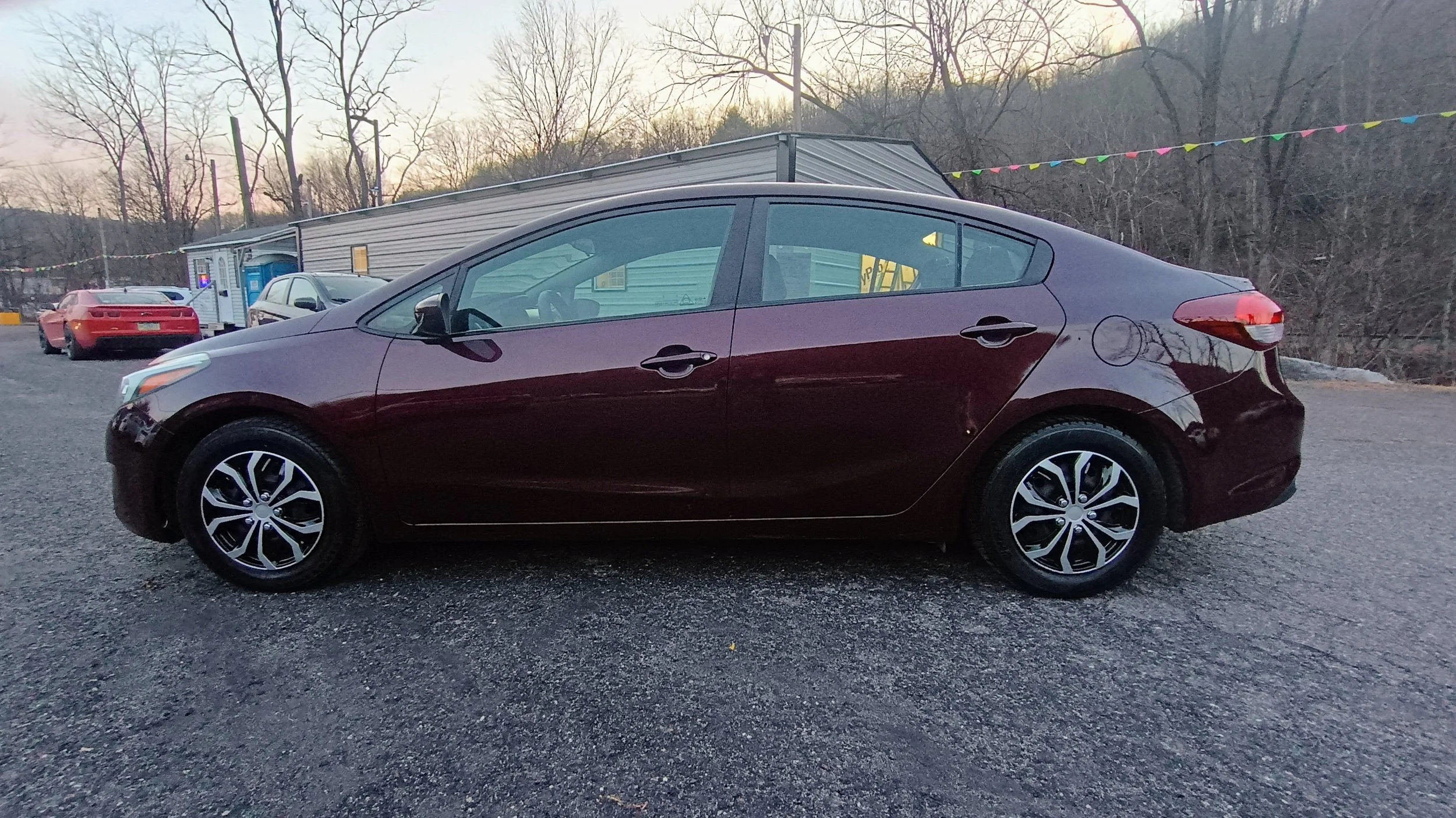 Side view of a dark red sedan parked on a gravel lot with other cars and a building in the background, during dusk.