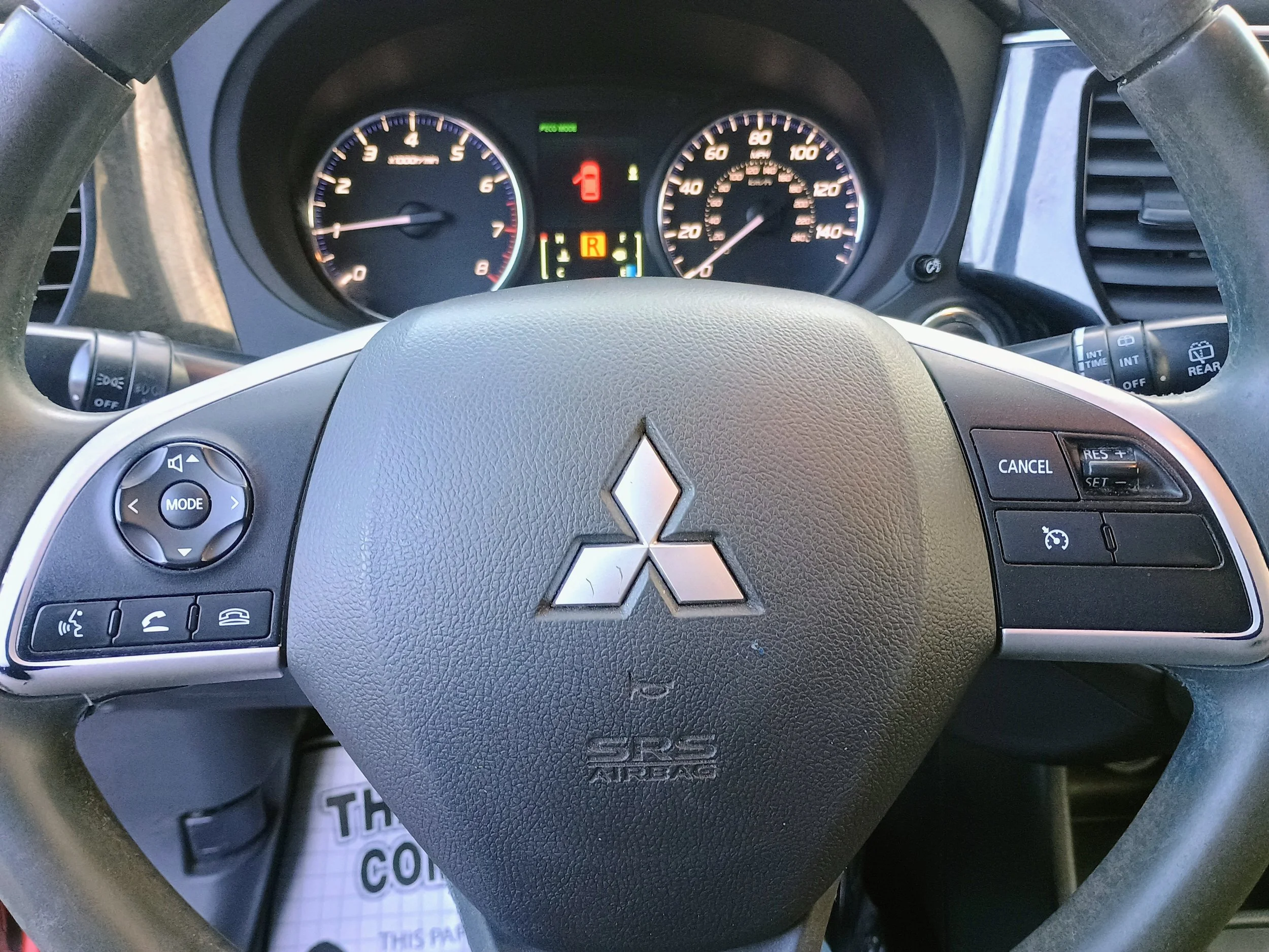 Dashboard view of a Mitsubishi vehicle steering wheel with various controls visible, including cruise control, phone, and mode buttons, and the speedometer and tachometer gauges in the background.