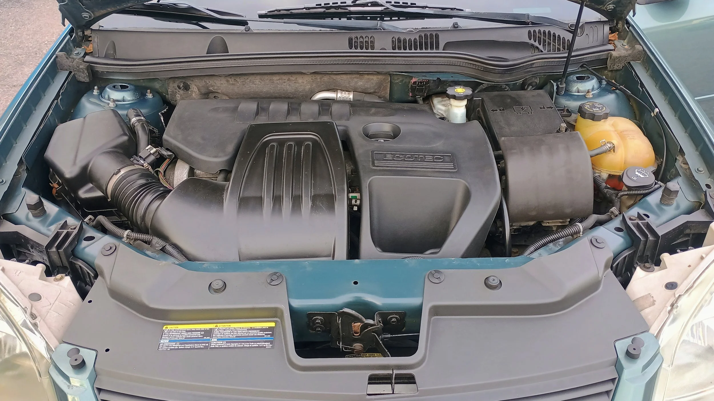Car engine bay showing various engine components, including the air intake, radiator cap, and coolant reservoir.