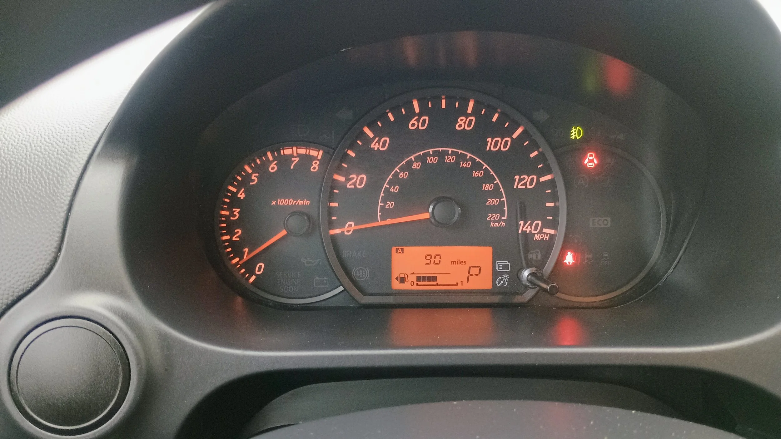 Car dashboard showing the speedometer, tachometer, battery, oil, seatbelt, and warning lights, with a digital display indicating 90 miles range.