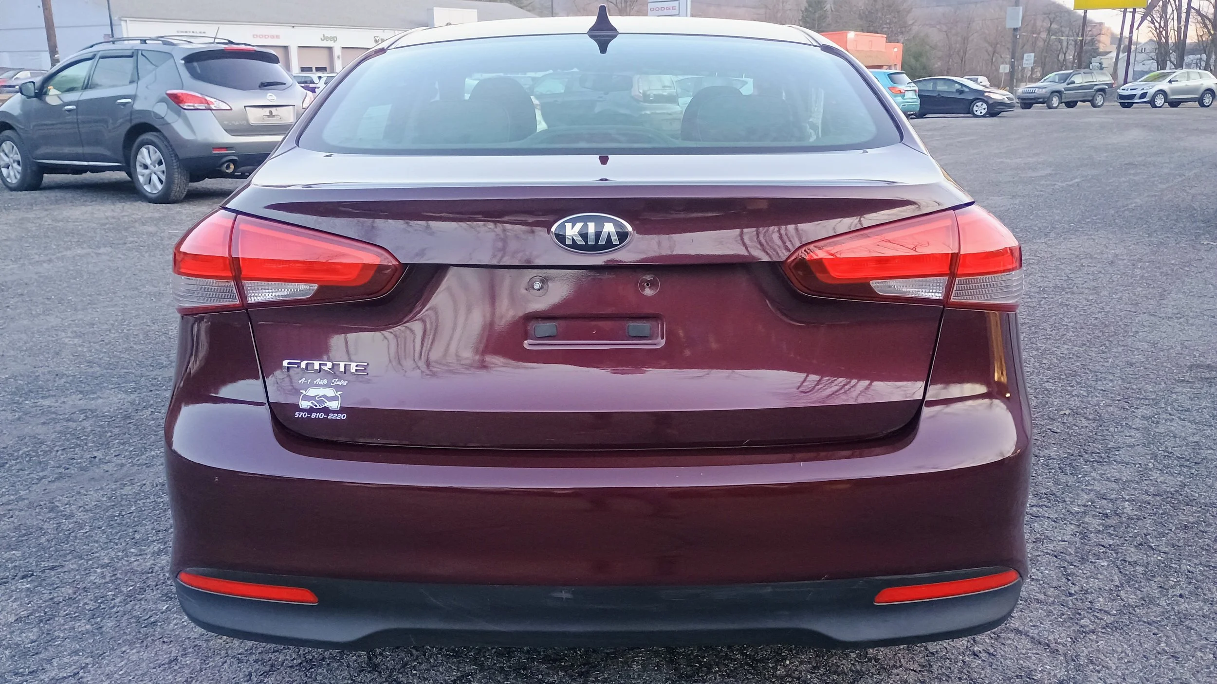 Rear view of a burgundy Kia Forte sedan parked on a gravel lot, with a dealership building in the background and other cars parked around.