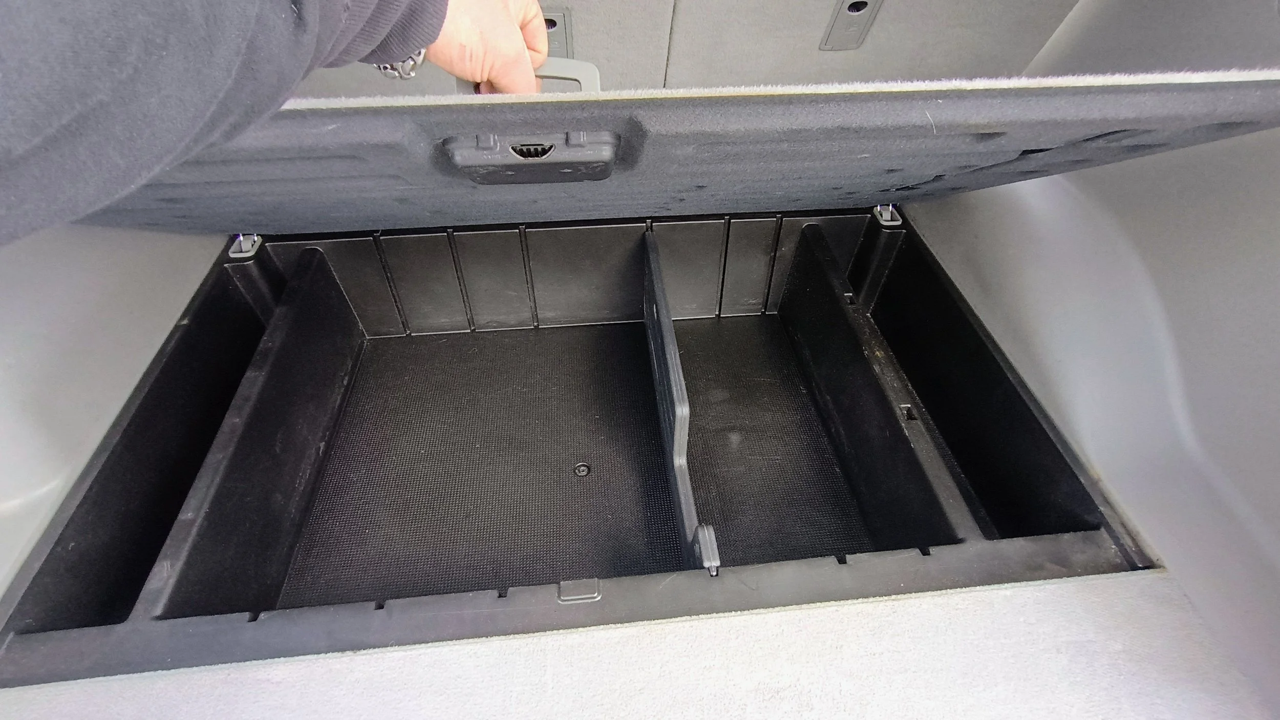 Under-seat storage compartment with a person opening the lid, showing empty divided sections inside.