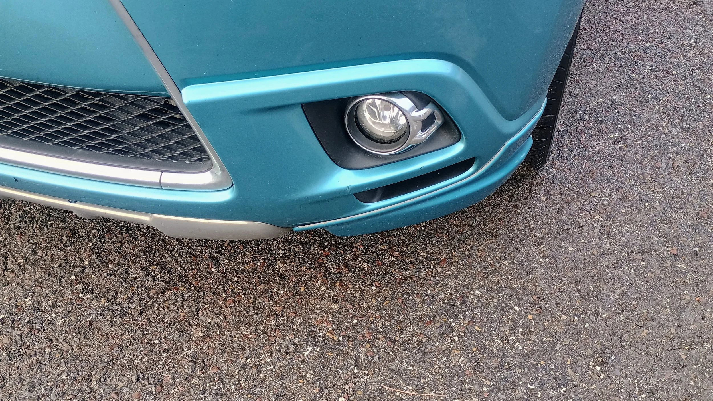 Close-up of the front right corner of a blue car, showing the fog light, grille, and tire on a gravel surface.