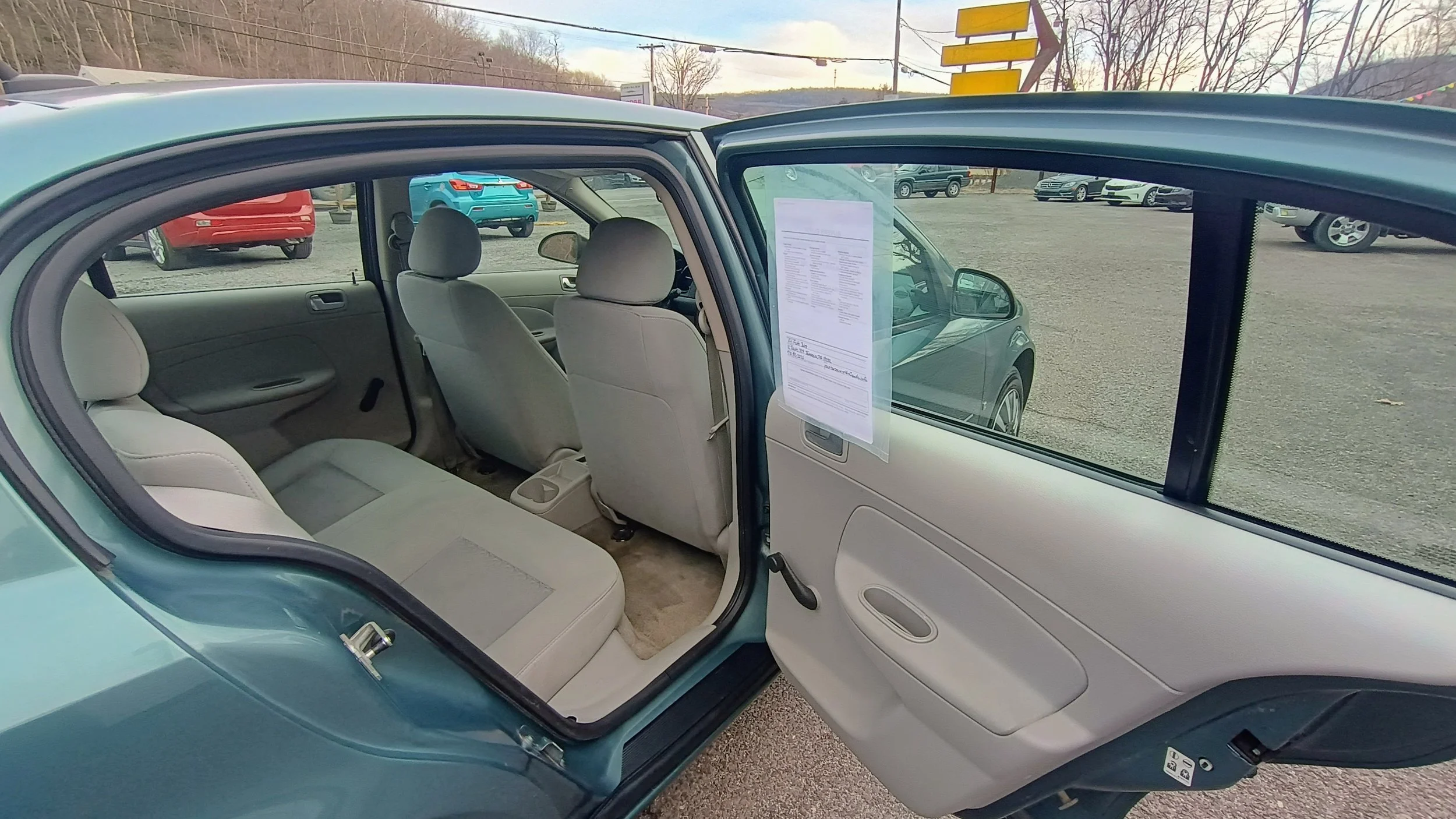 Inside view of a light blue compact car with beige interior, showing the open rear and front doors, with other cars in a dealership lot in the background.
