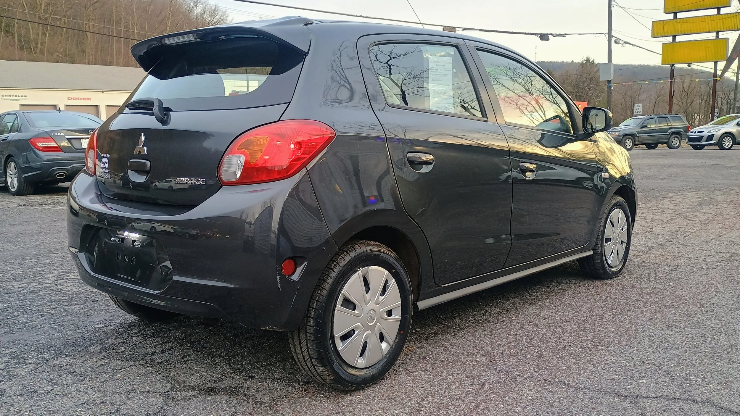 Black Mitsubishi Mirage compact hatchback car parked in a car dealership lot with other vehicles in the background, overcast sky.