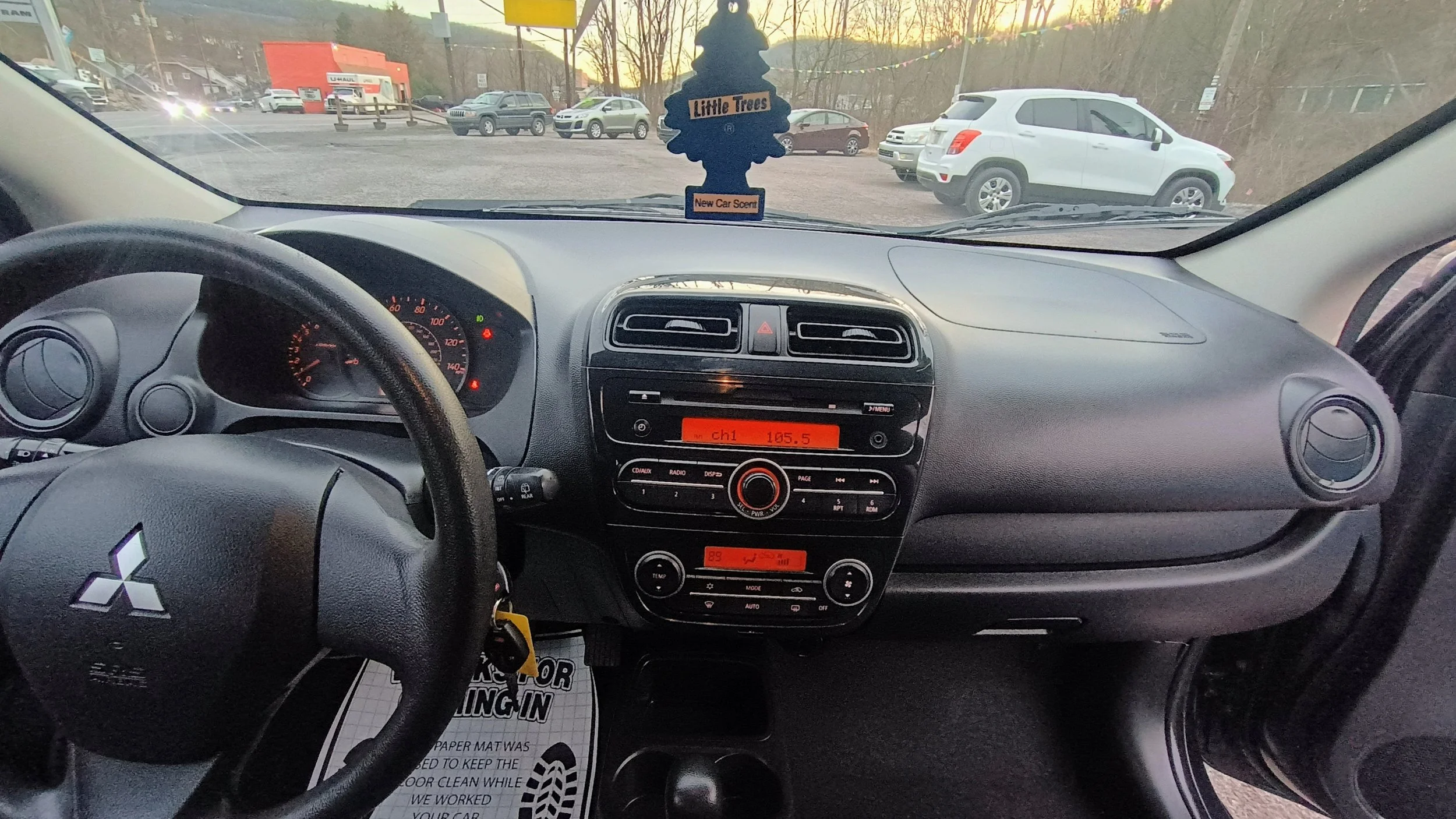 Inside a Mitsubishi car showing the dashboard, steering wheel, and a view through the windshield of a parking lot with several cars and a U-Haul building in the background during sunset. An air freshener shaped like a tree hangs from the rearview mirror.
