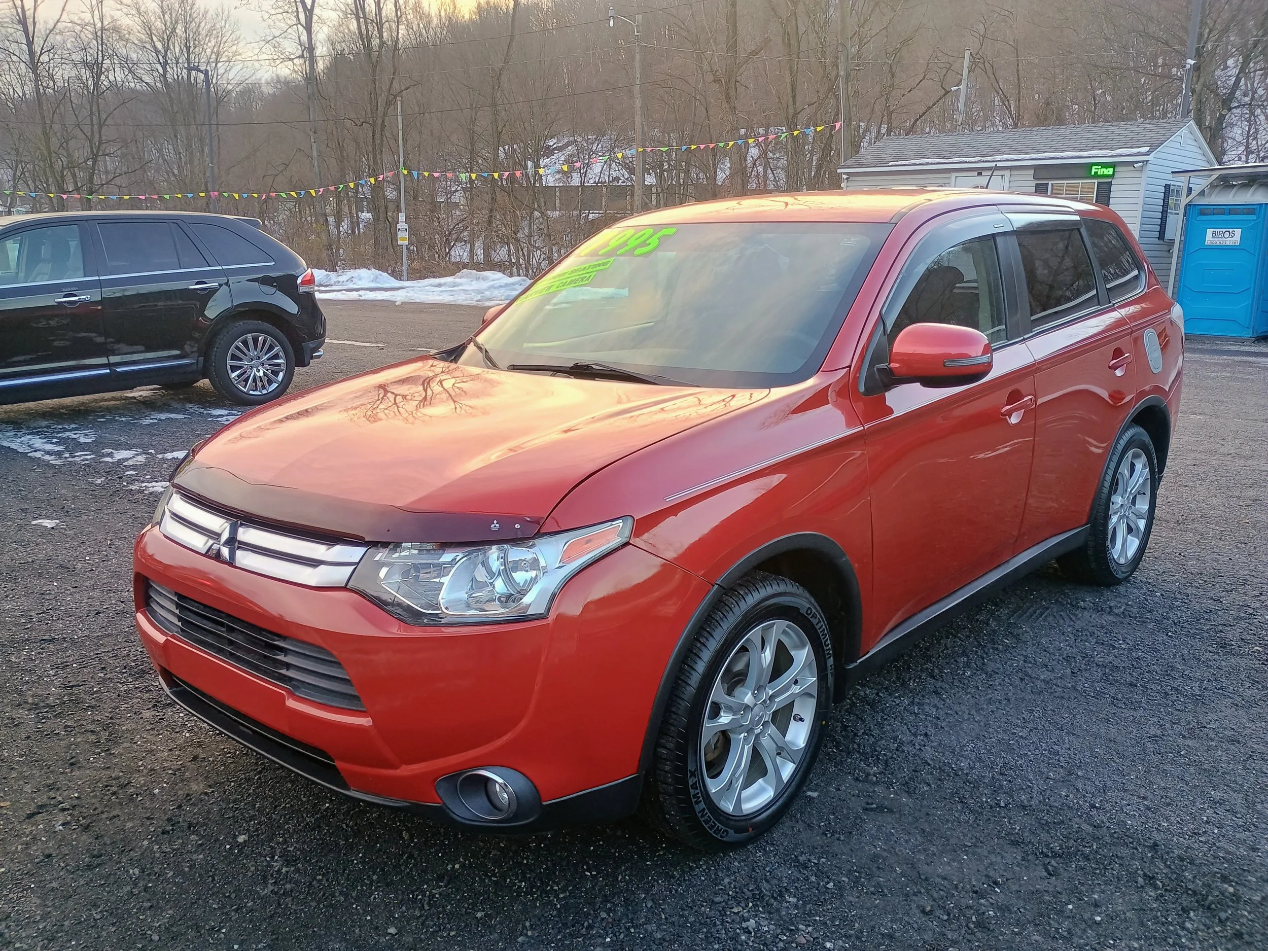 Red mid-sized hatchback car parked in a lot, with a green price tag on the windshield, surrounded by other vehicles, trees, and snow patches.