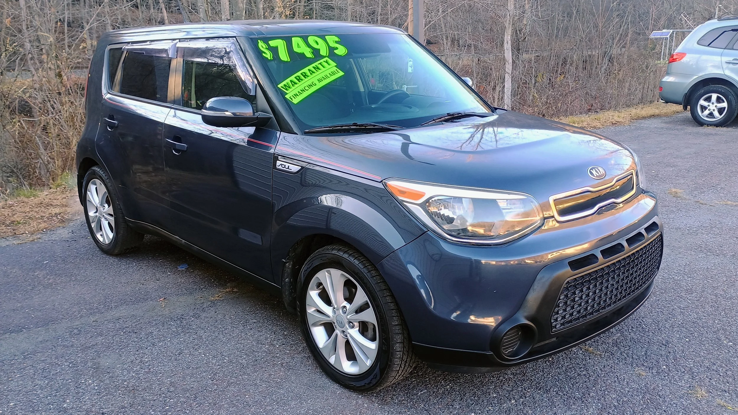 Dark blue Kia Soul parked in a car lot with a bright green price sign of $7495 on the windshield and a yellow warranty sign, with other vehicles and trees in the background.