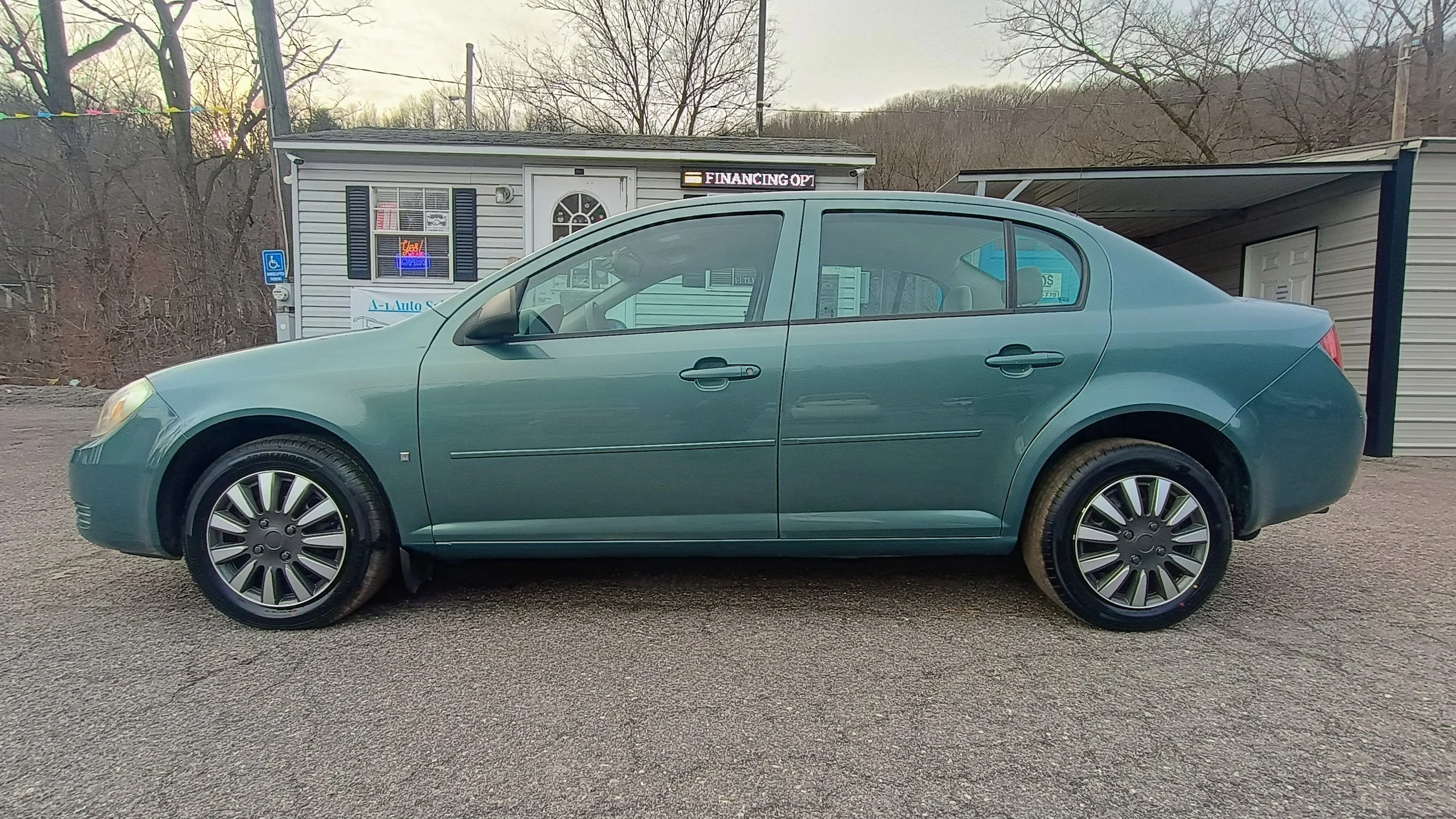 Side view of a teal-colored sedan parked on a gravel lot in front of a small building with a sign that reads 'Financing Op' and a neon 'Open' sign in the window.