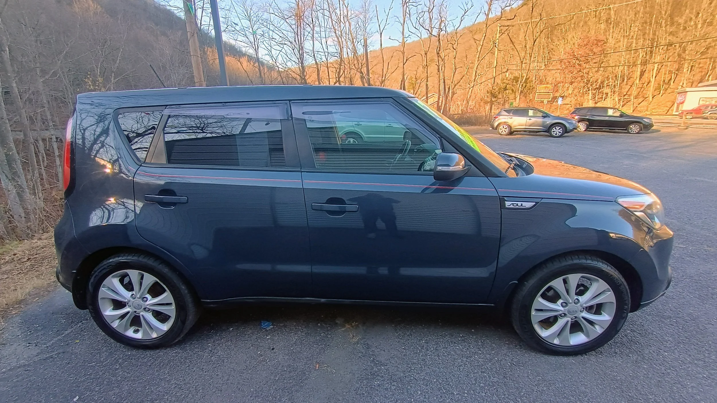 Side view of a dark blue Kia Soul parked in a gravel lot with other cars in the background, trees, and hills during sunset.