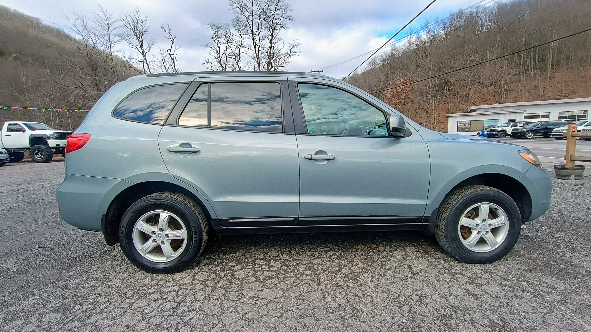 A light blue SUV parked on a gravel lot with a mountain and some trees in the background. Several other vehicles are visible, including trucks and cars, near a white building with large windows.