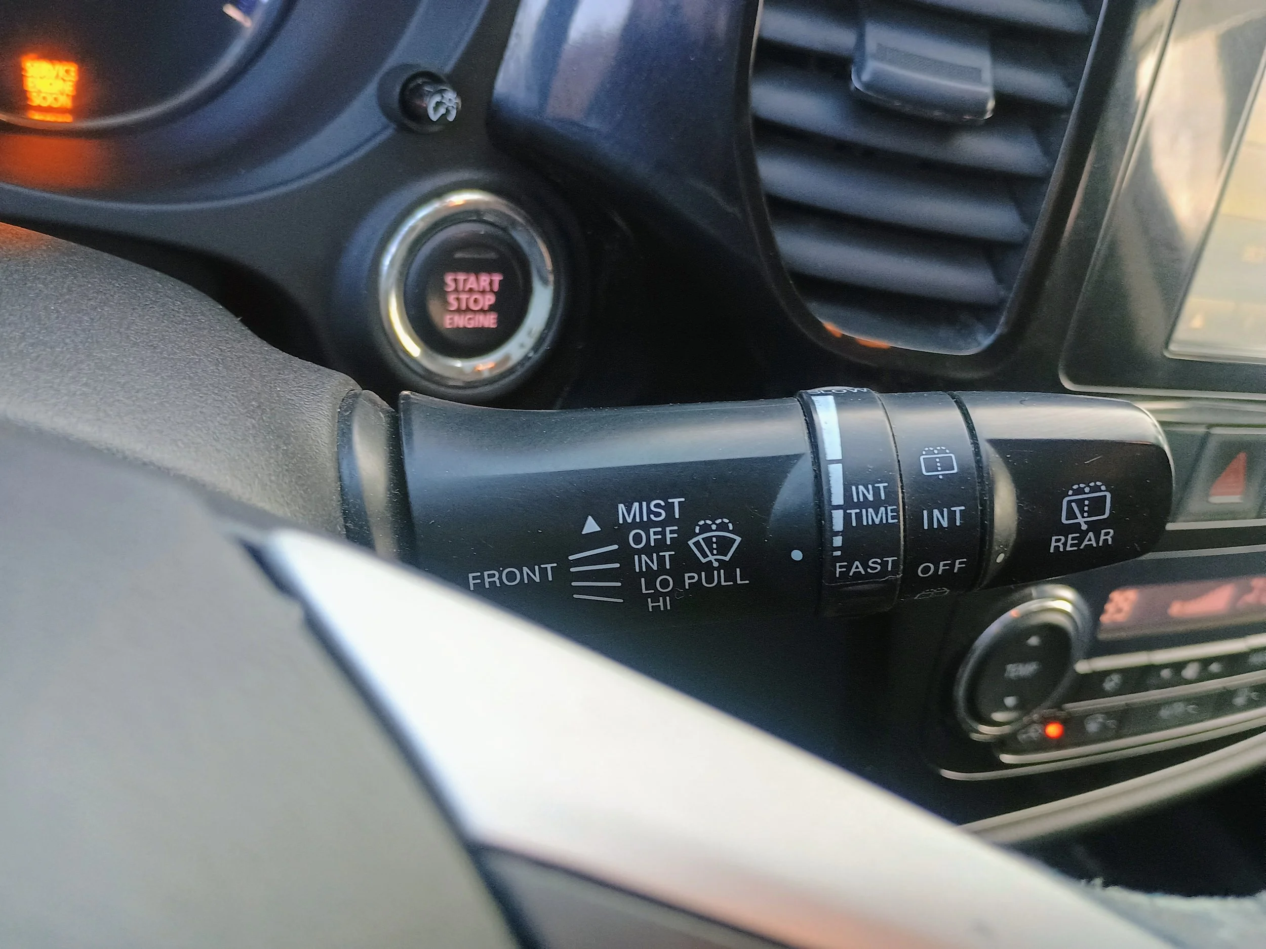 Close-up of a car's windshield wiper control stalk with various settings and icons, including front and rear wipers, intermittent, and mist functions. The dashboard and engine start/stop button are partially visible in the background.