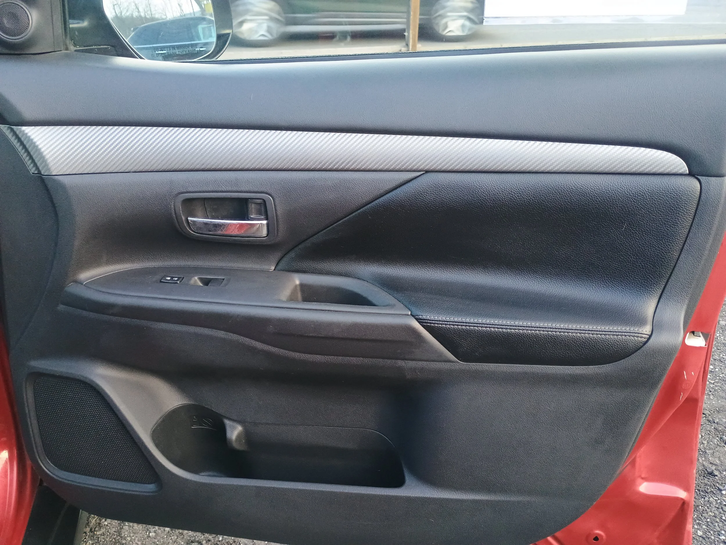 Inside view of a car door with a black and silver handle, window control, and speaker, with a red exterior visible at the bottom.