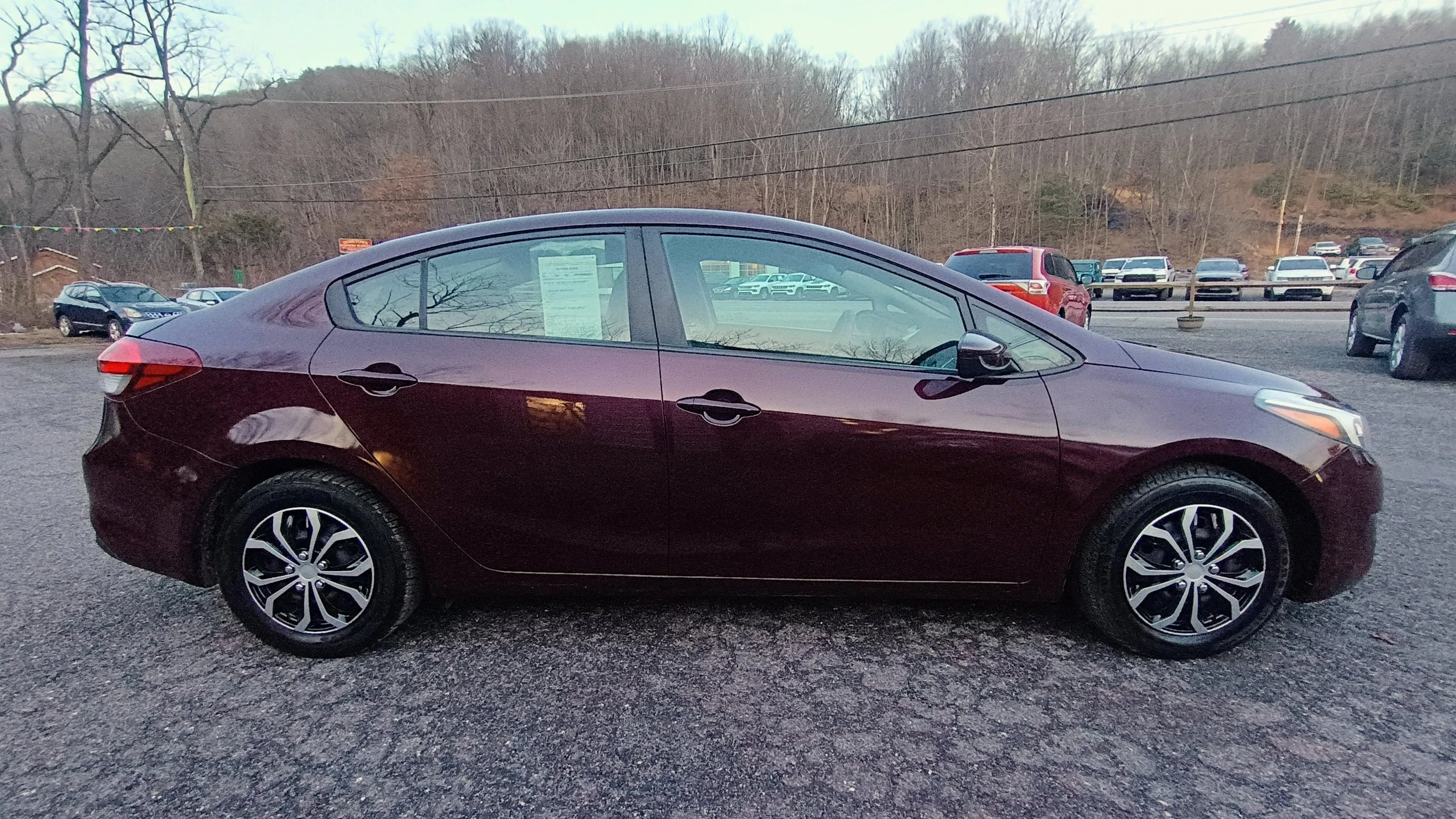 A maroon four-door sedan parked on a gravel lot with other vehicles visible in the background, and bare trees on a hill behind.