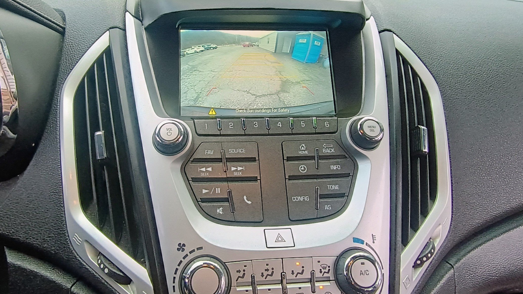 The image shows a car's dashboard with an active rearview camera display. The screen displays the view behind the vehicle with parking guidelines. The dashboard has various buttons and controls for audio, navigation, and climate settings.