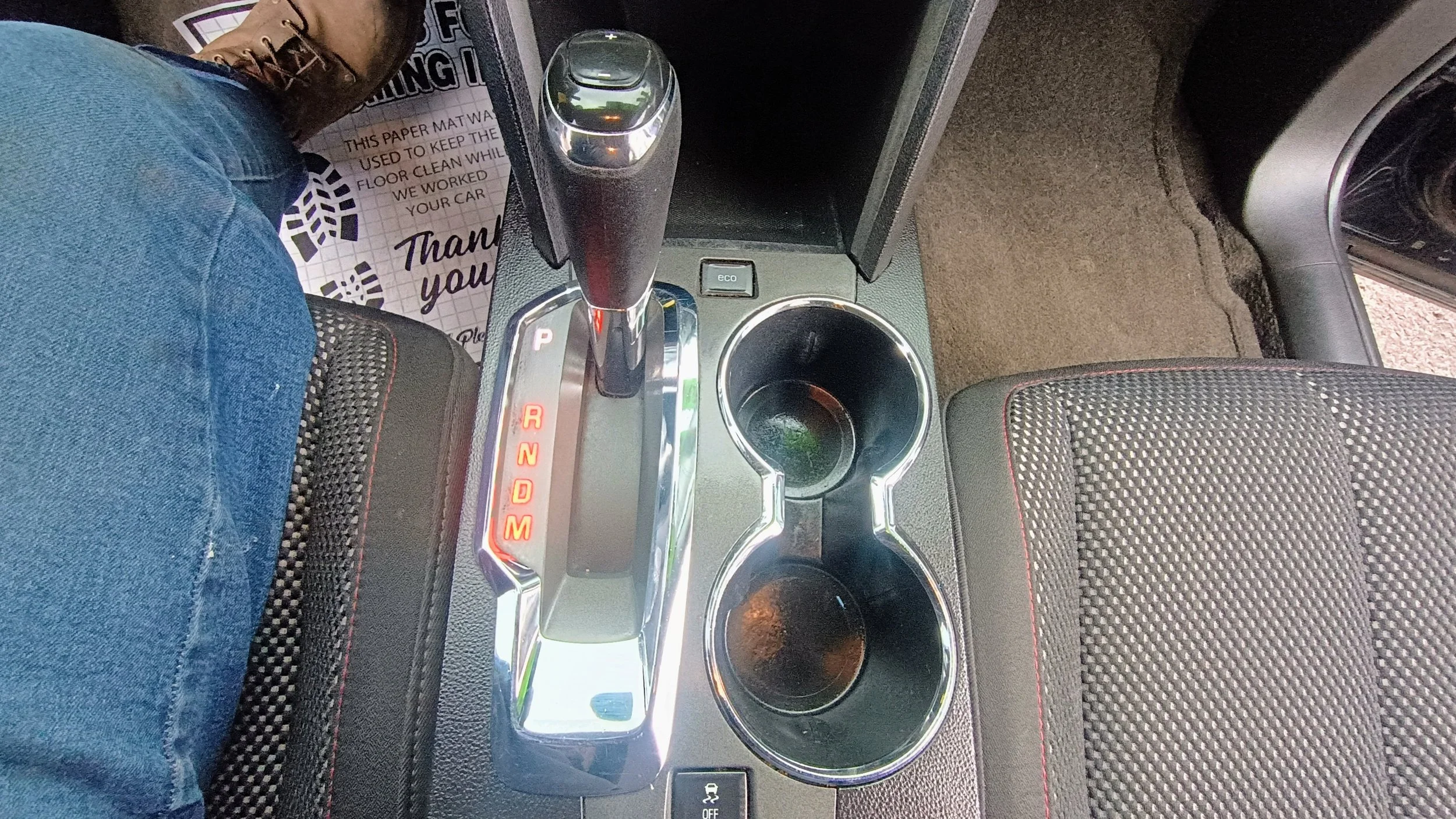 Interior view of a car's center console with gear shift, two cup holders, an 'eco' button, and part of the driver's leg wearing a boot.