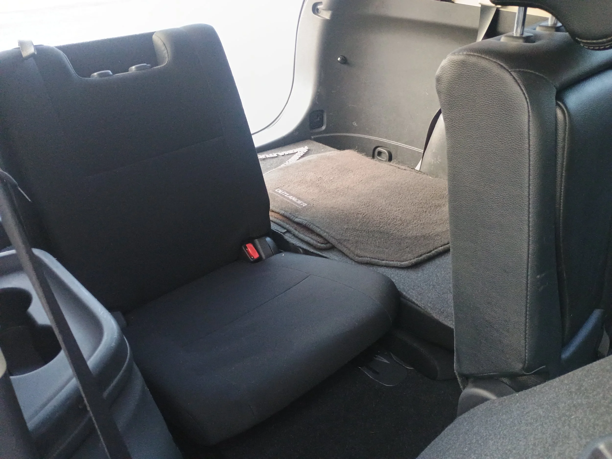 Interior of a vehicle showing the backseat with a folded down seat back and a blanket on top, with black upholstery and a gray carpeted floor.