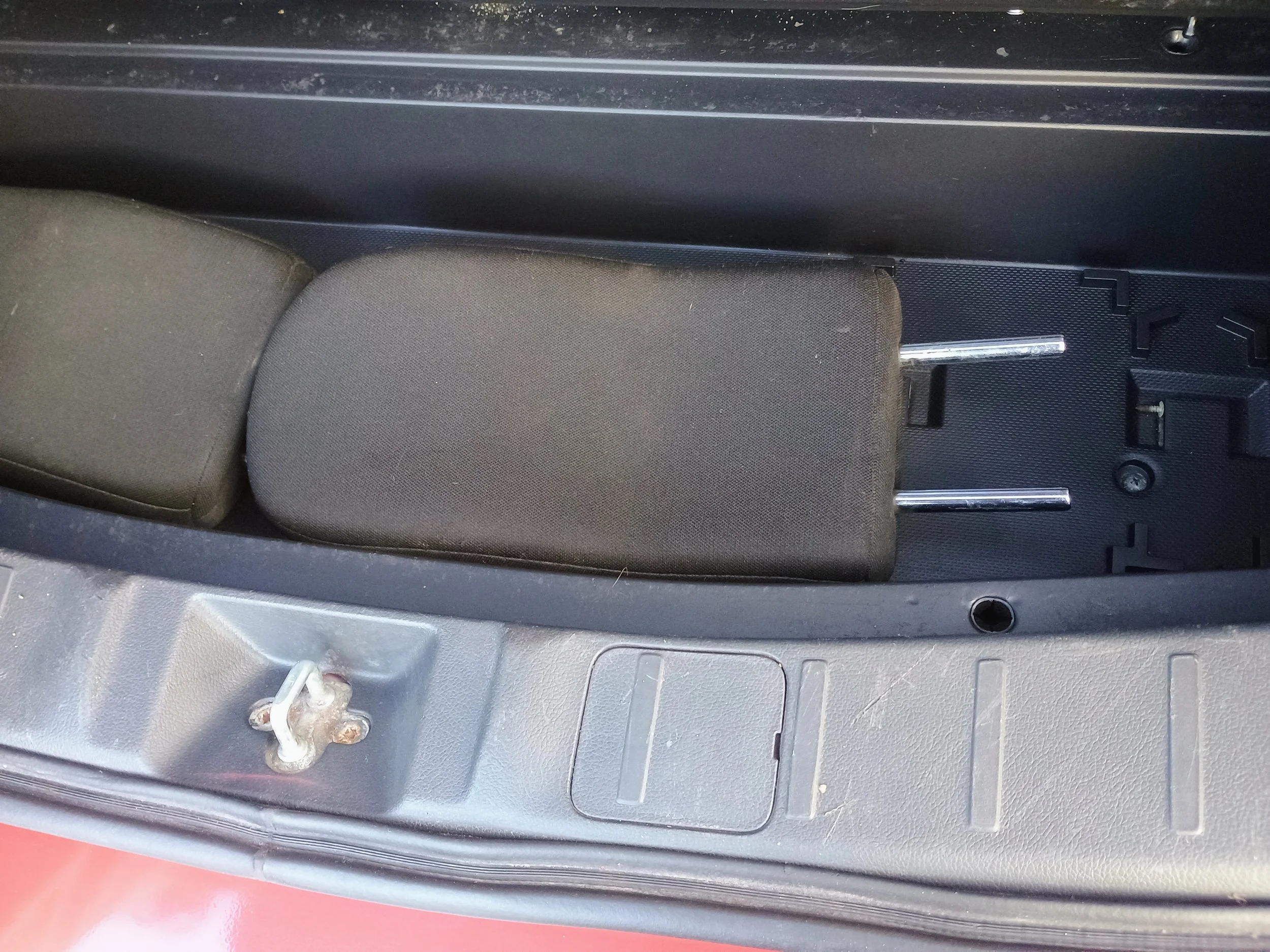 The image shows the interior of a vehicle's trunk area with a black padded headrest or cushion, with a metal support rod, and part of a gray plastic panel with a hook and small hatch or compartment.