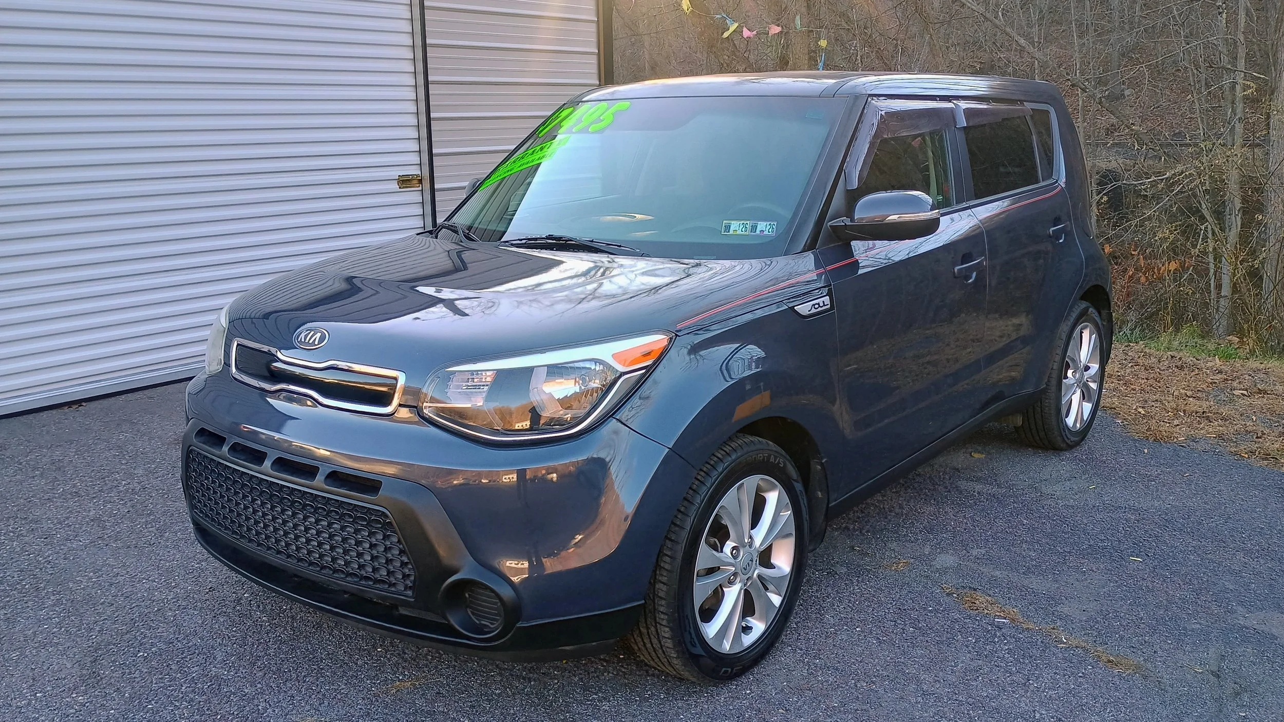 A blue Kia Soul compact car parked outdoors next to a metal garage door, with a reflection of the sky on its hood and windshield, and a price sticker on the windshield.