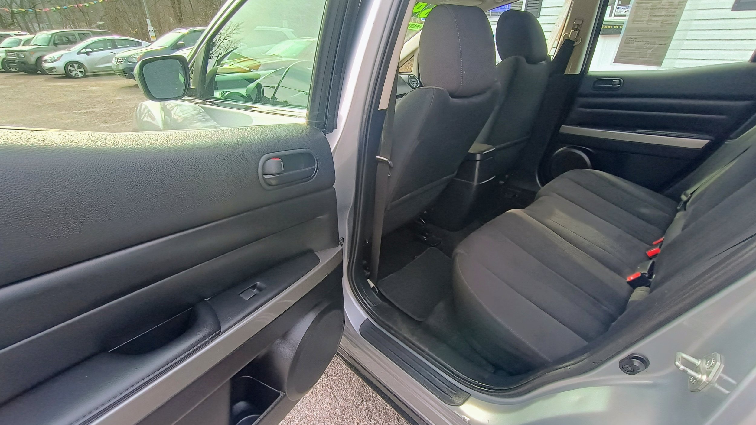 Interior view of a vehicle showing the back seats and part of the front seats, with the driver-side door open, parked outside in a lot with other cars visible in the background.