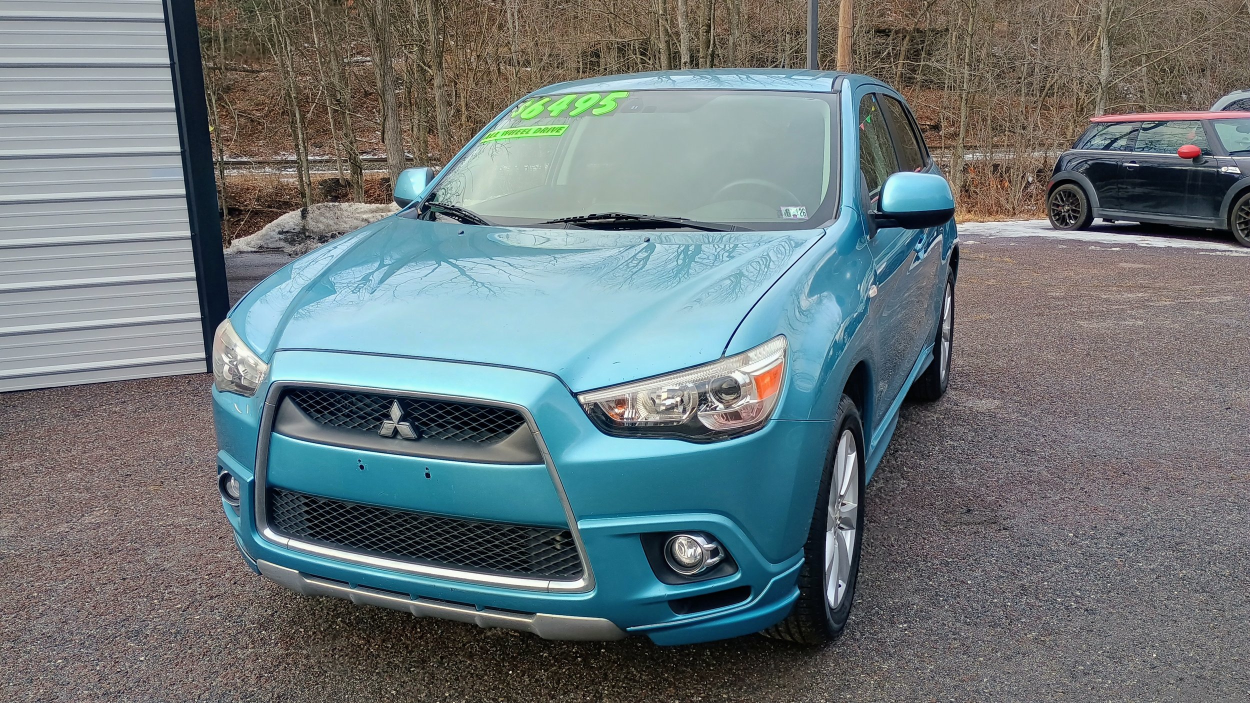 Blue Mitsubishi SUV parked in a lot during daytime with trees and a small hill in the background, and a sign on windshield showing a price of $5,495.