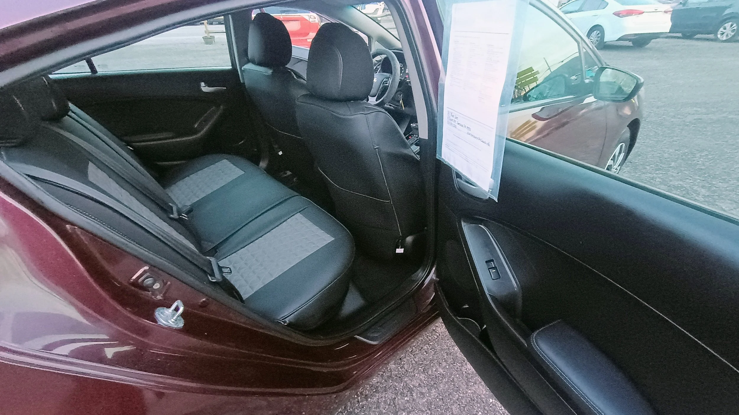 Interior of a car, showing the backseat area, with open front passenger door, black and gray seats, and a window sticker on the windshield. Other cars are visible parked outside.