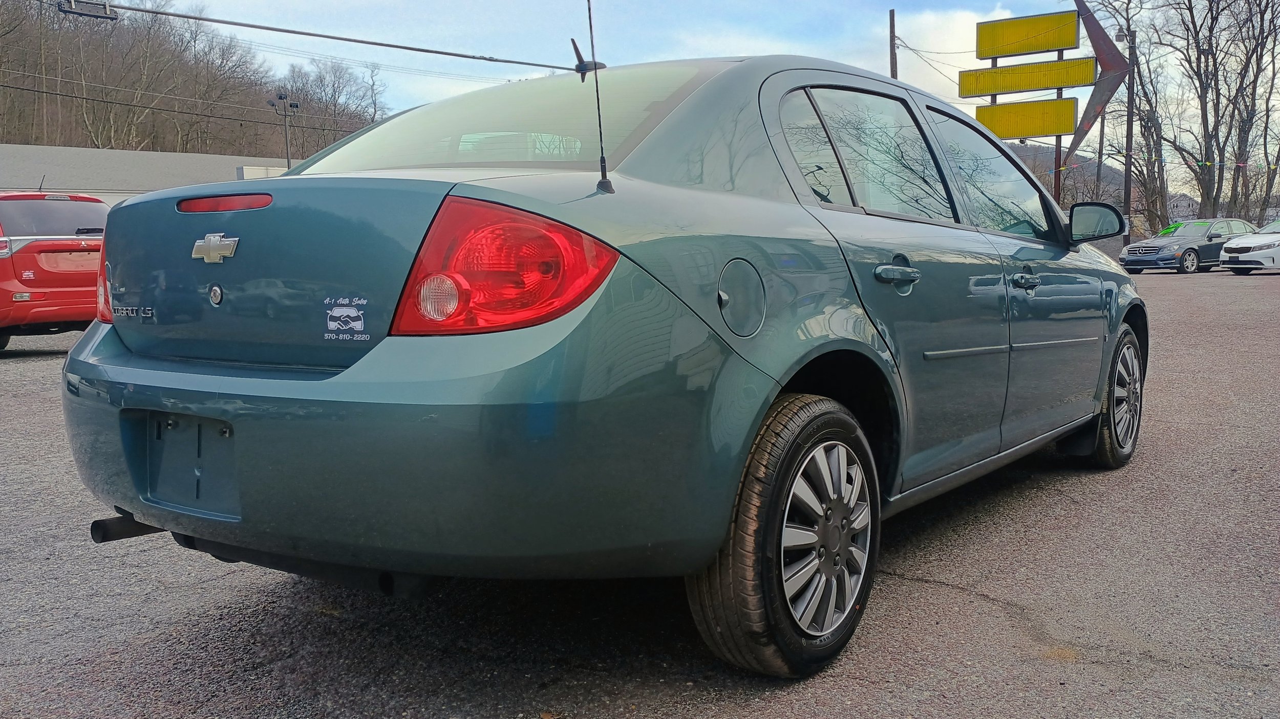 Rear and side view of a teal Chevrolet Malibu LS sedan parked on an asphalt lot, with other cars and a yellow directional sign in the background.