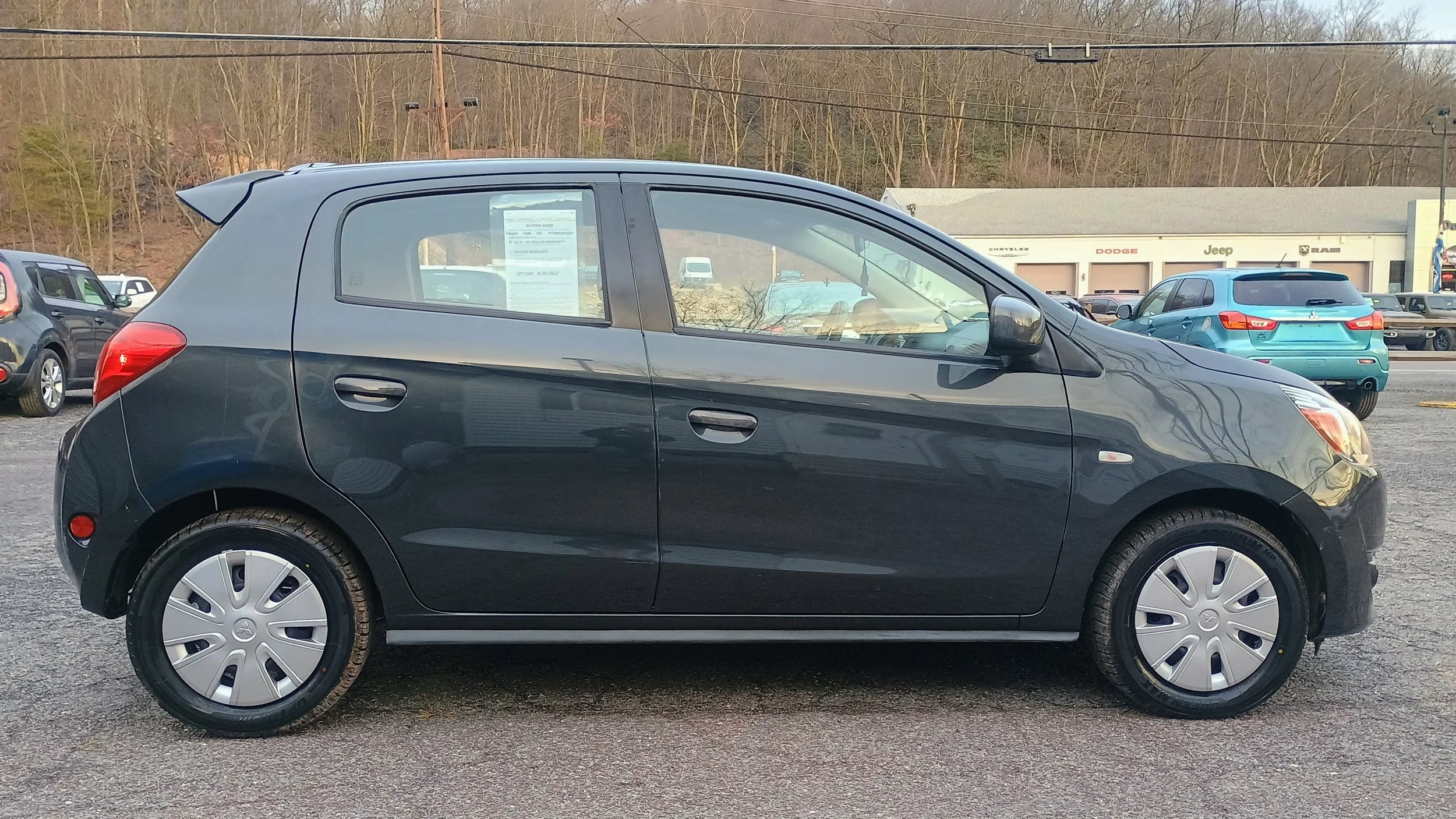 A black compact hatchback car parked on a dealership lot, with other cars visible in the background and a dealership building with various brand logos.