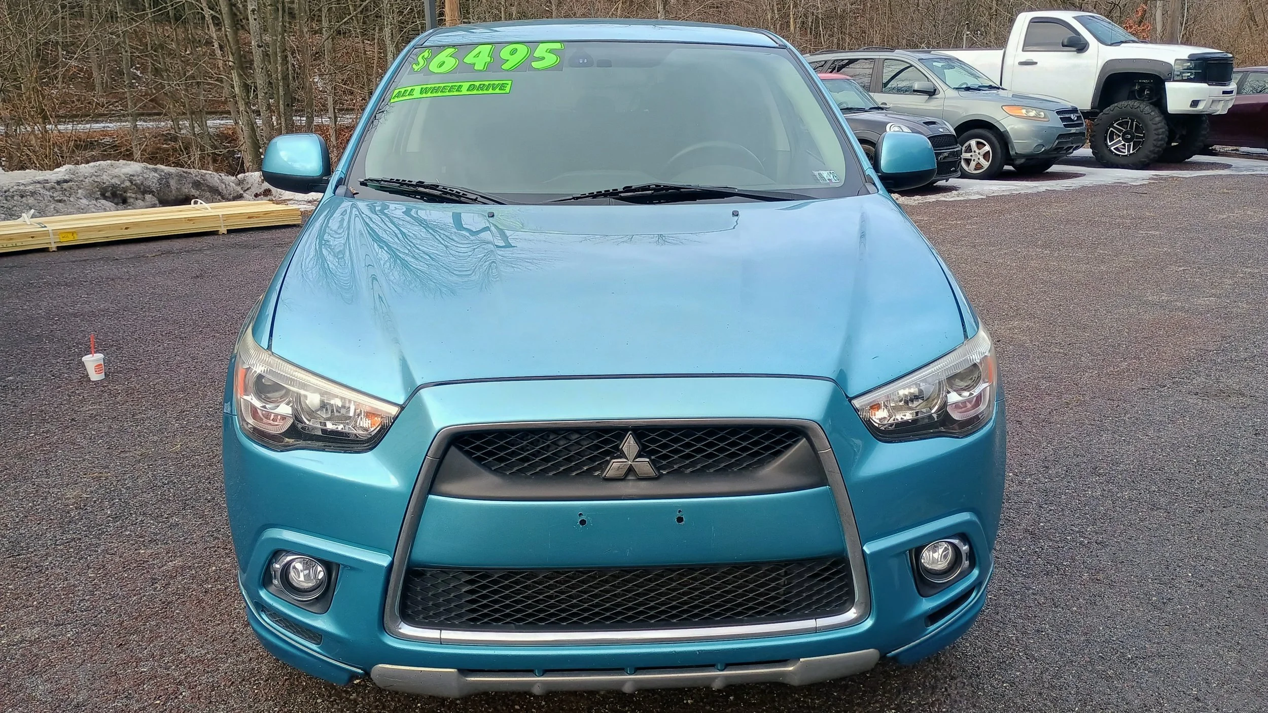 Front view of a blue Mitsubishi sedan parked on a gravel lot with green price and details on windshield, and other vehicles in the background.