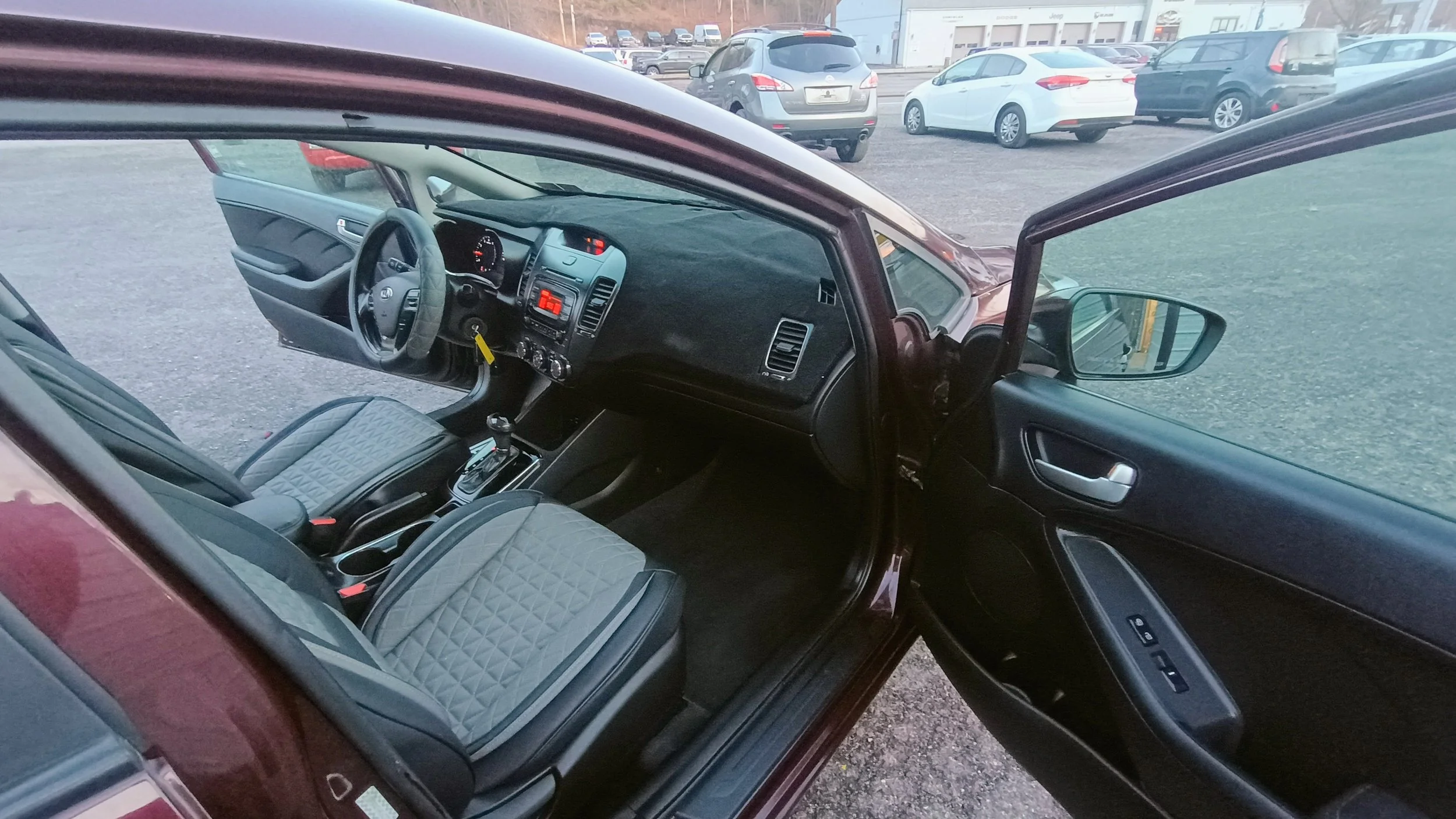 Inside view of a car with gray quilted seats, black dashboard, and gear shift. Open door showing side mirror and window controls.