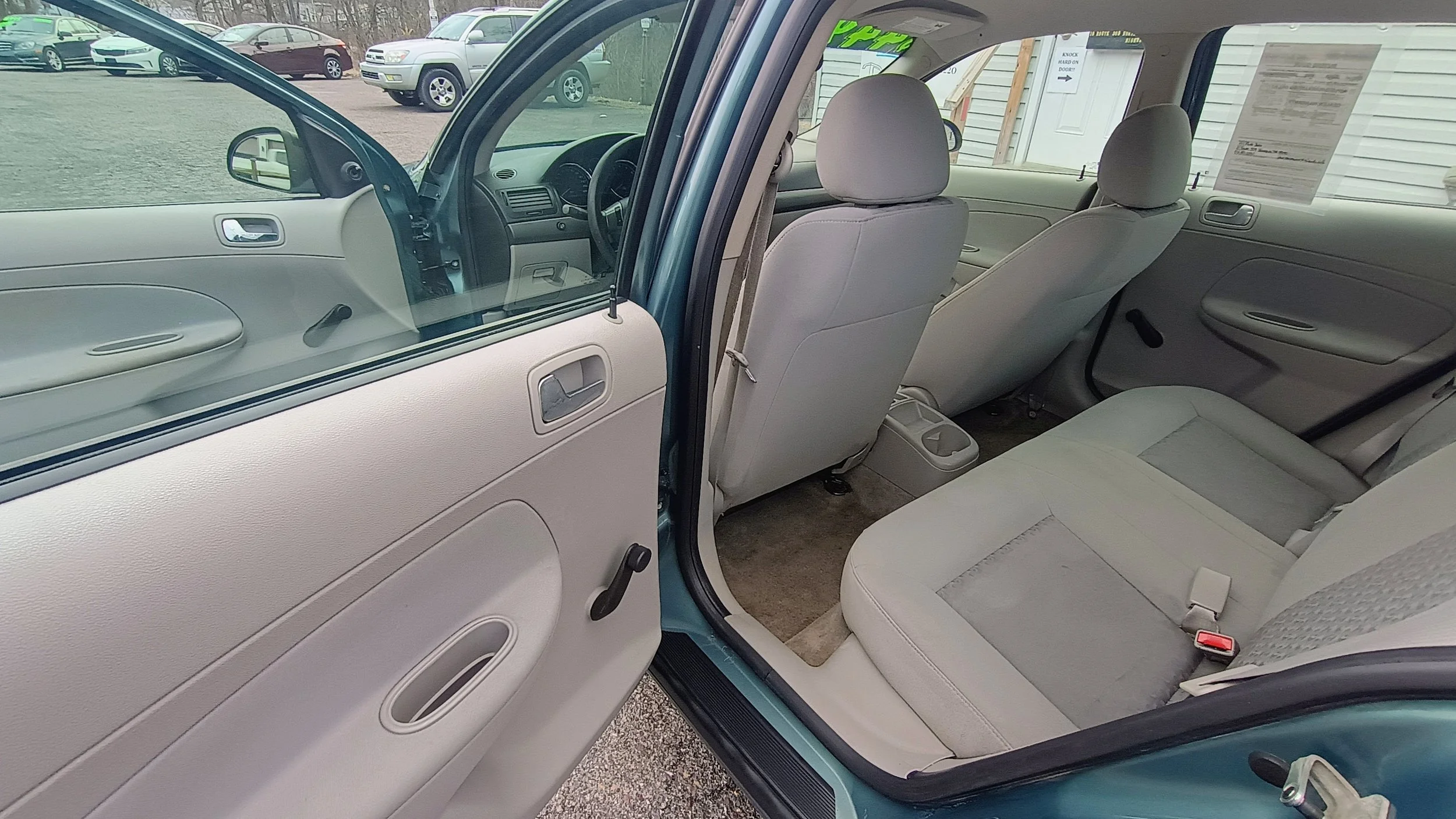 Interior of a light-colored sedan showing the backseat area with beige fabric seats and visible seatbelt, view through an open rear passenger door.