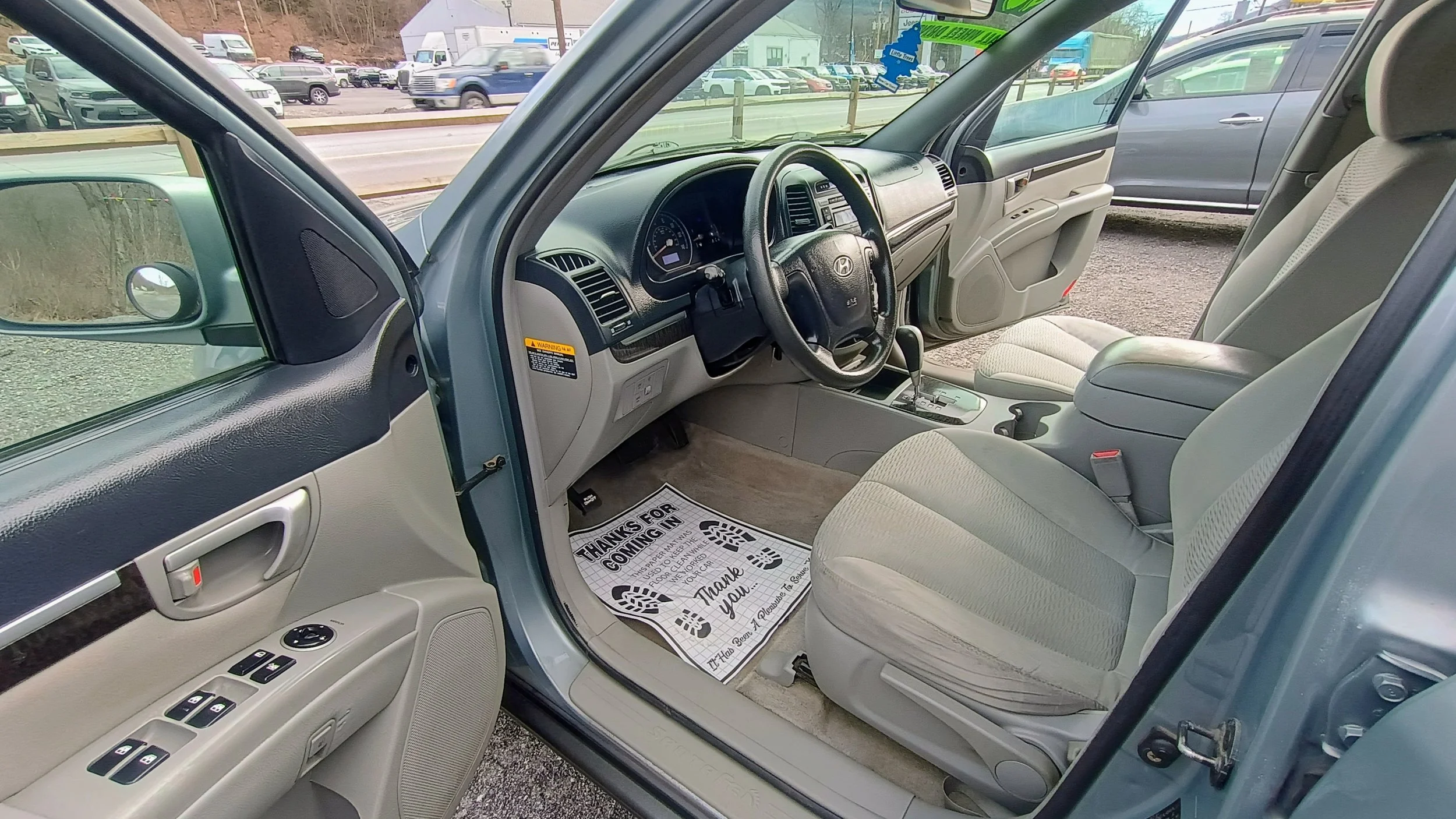 Interior of a silver Toyota SUV showing the driver's side with the door open, gray fabric seats, a black dashboard, and a gear shift. There is a paper floor mat that says 'Thanks for coming in' on the driver’s side.