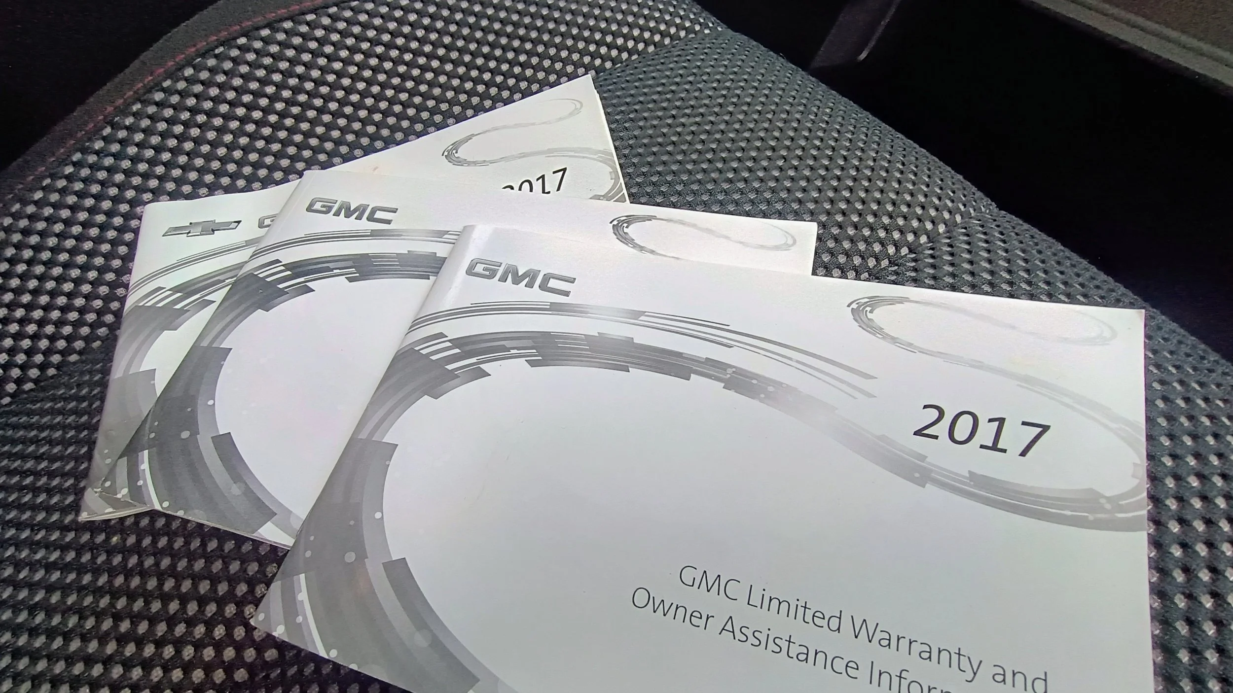 Three white booklets or brochures with gray graphics and 'GMC' logo, titled '2017,' placed on a black textured car seat.