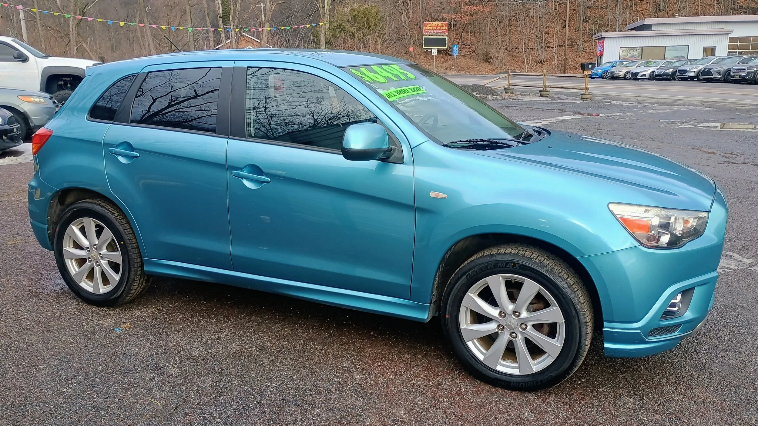 Blue compact hatchback car for sale at a used car dealership with a bright green sign on the windshield displaying the price and sale information.