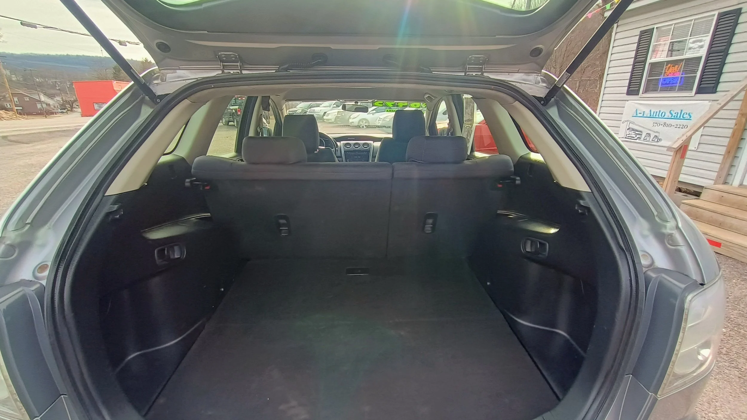 Rear view of an open SUV trunk showing a spacious cargo area with third-row seats up in a parking lot near a building with a sign that says 'A-L Auto Sales'.