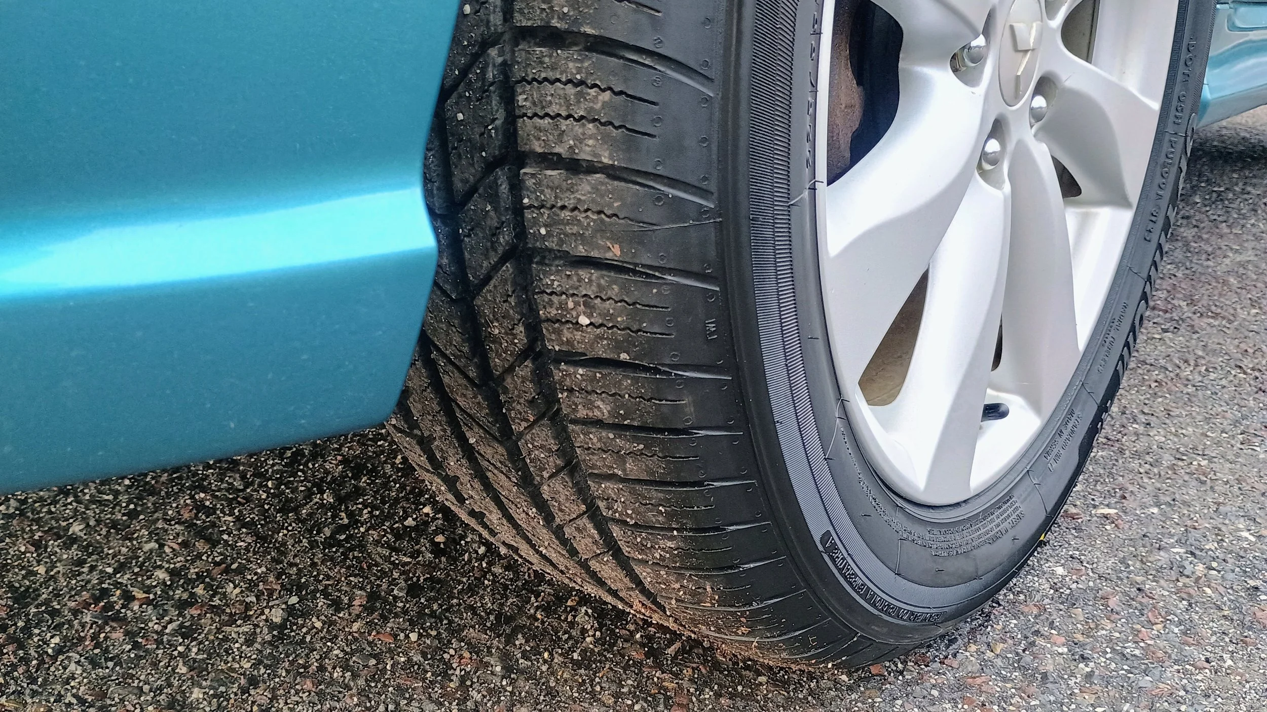 Close-up of a car tire with tread pattern, mounted on a white metal rim next to a teal-colored car body, on a gravel surface.