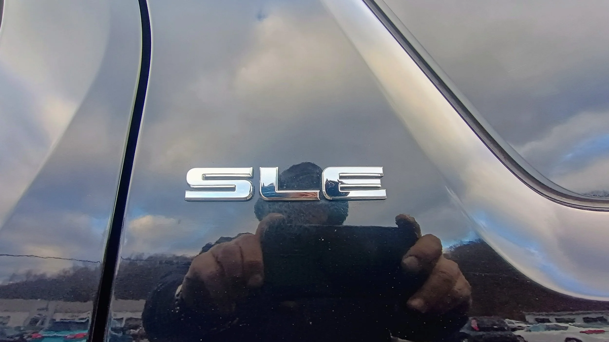 Close-up of a car's rear with a chrome 'SLE' emblem, reflecting the sky, clouds, and the person taking the photo.