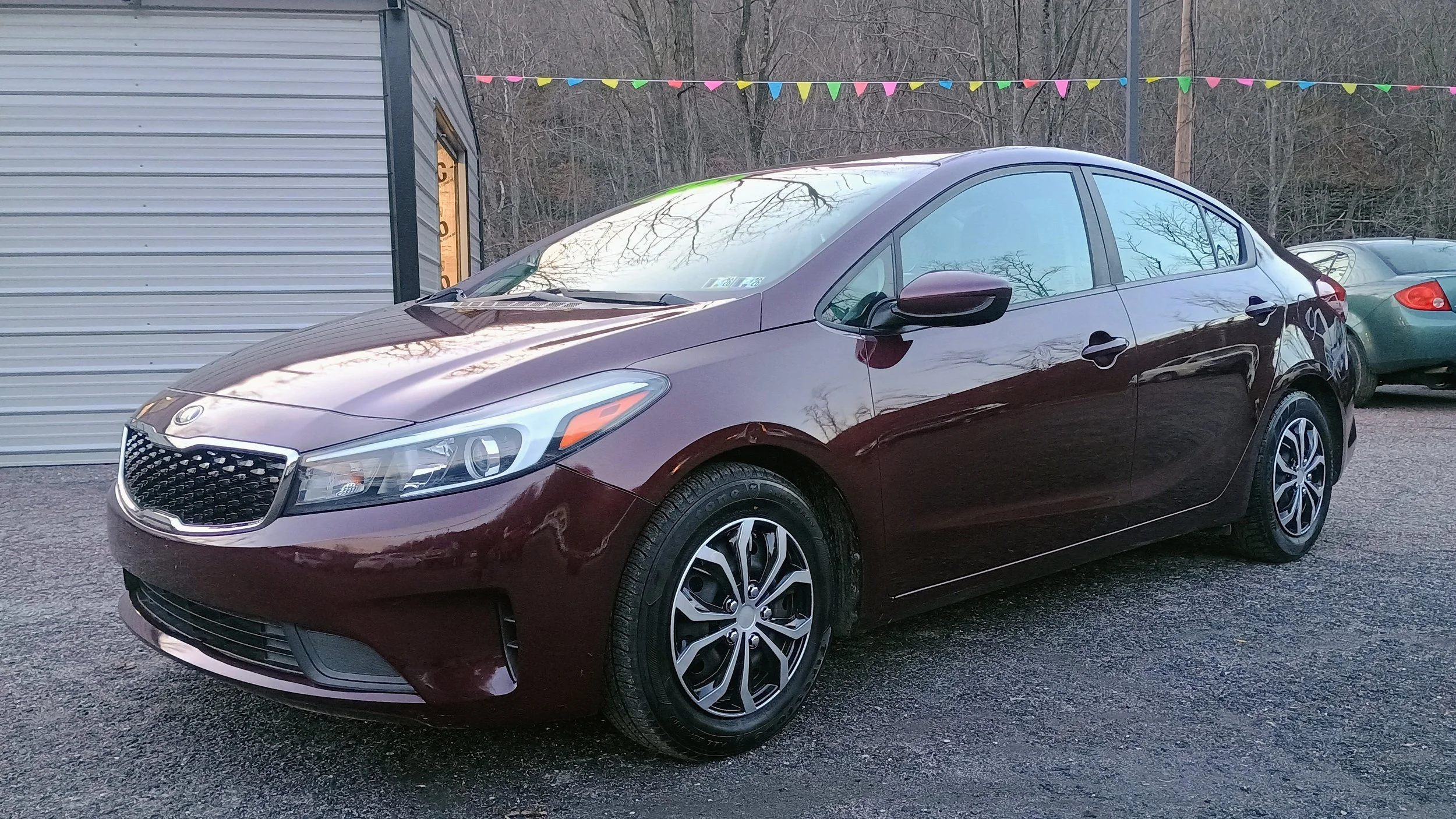 A maroon compact sedan car parked on a gravel lot with buildings and other cars in the background, and colorful triangular flags hanging overhead.