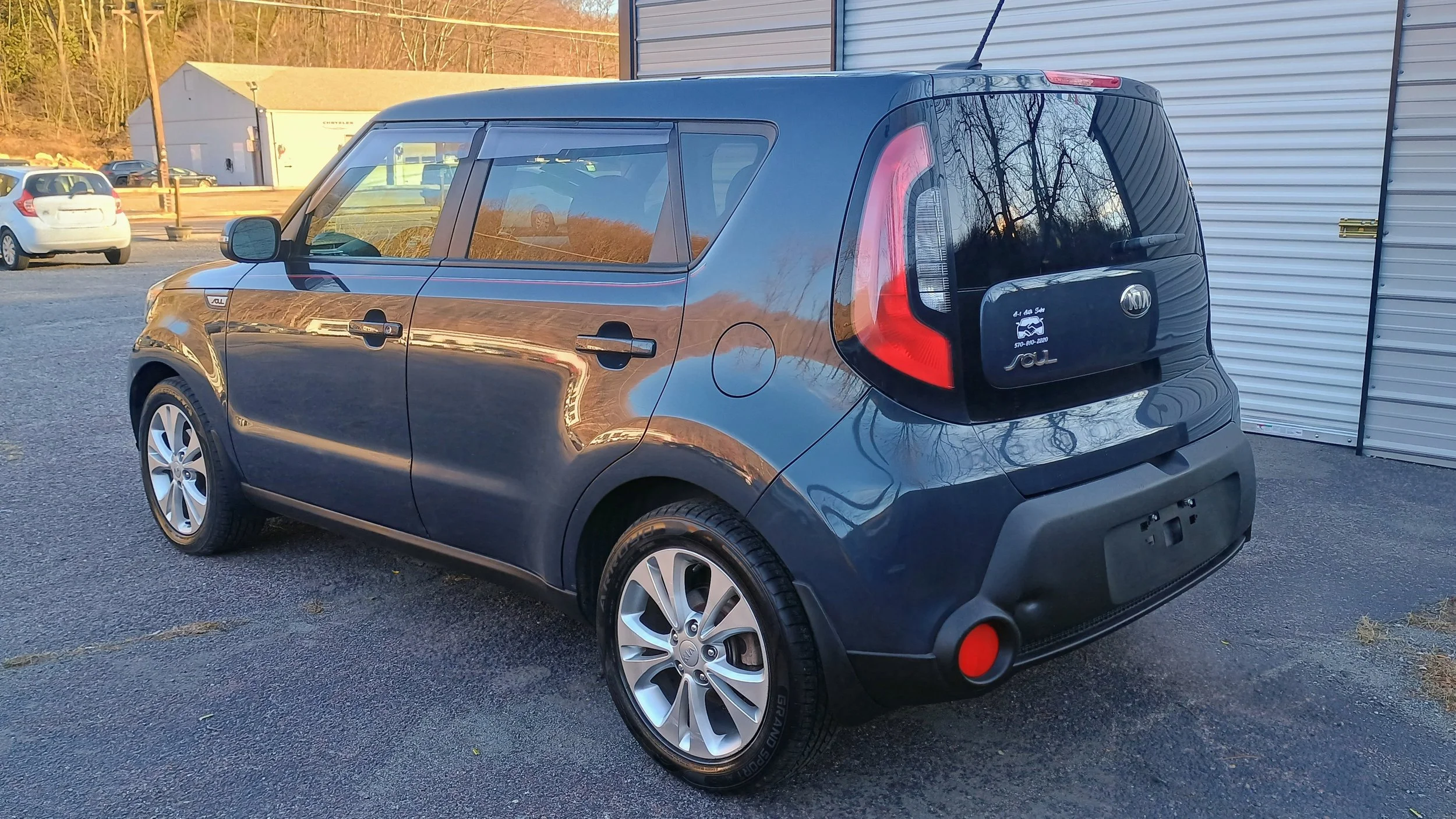 Black Kia Soul parked on asphalt at a car dealership lot with other cars and a building in the background.