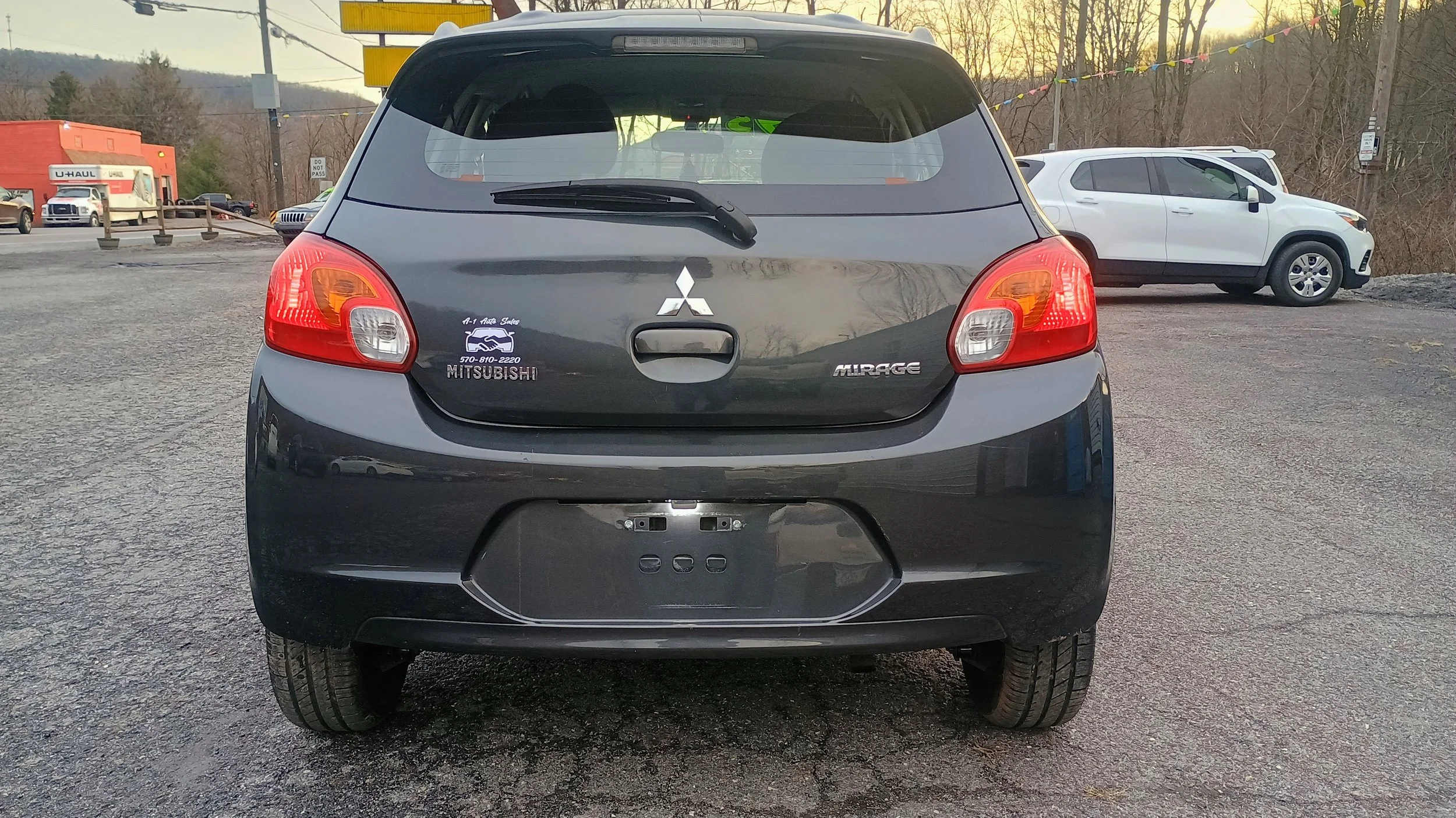 Rear view of a black Mitsubishi Mirage hatchback with a dealership sticker and no license plate, parked in a lot with other vehicles and trees in the background.