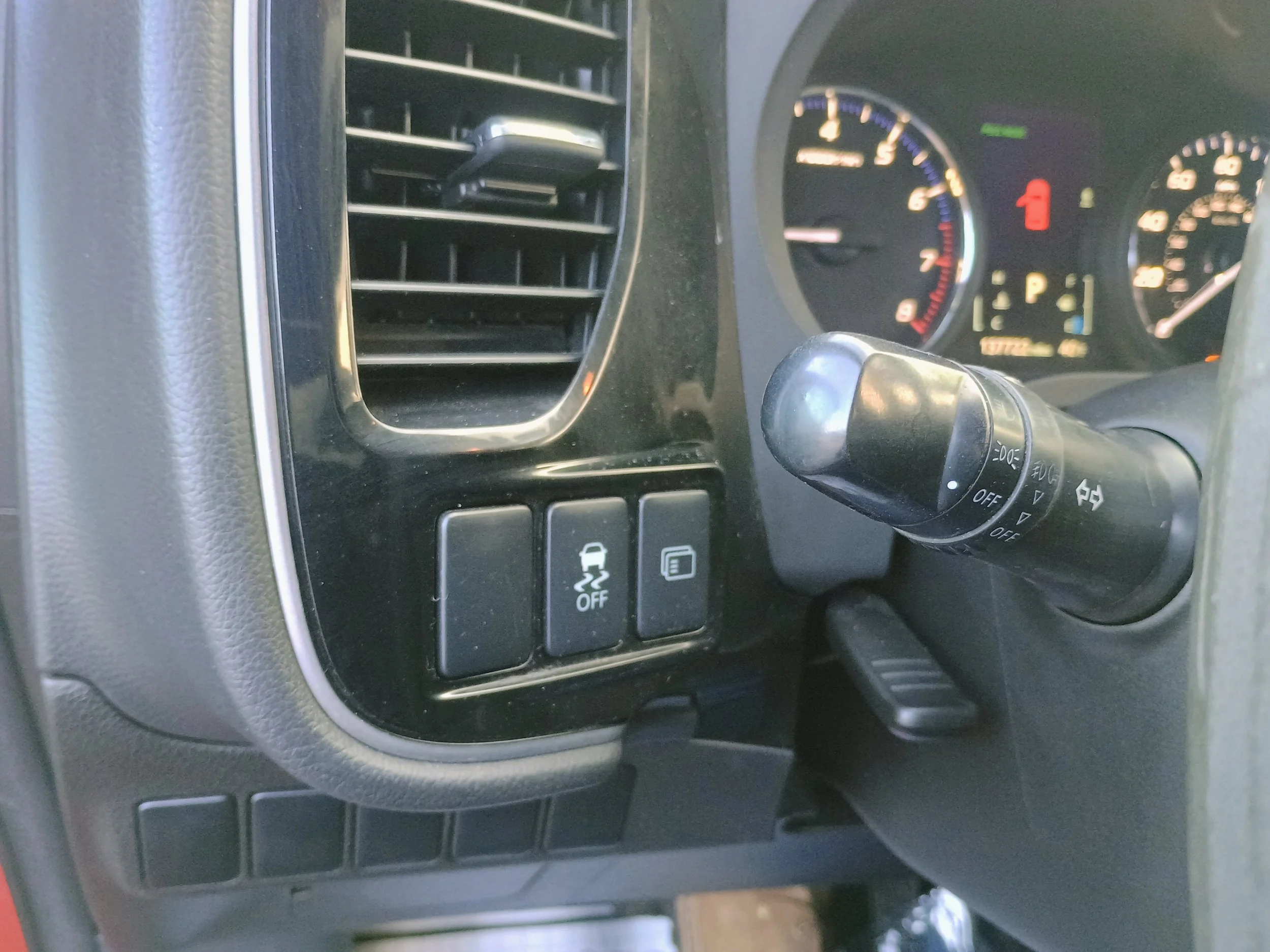 Close-up of a car dashboard showing an air vent, control buttons including one for traction control, and part of the steering wheel with turn signal and headlight controls.