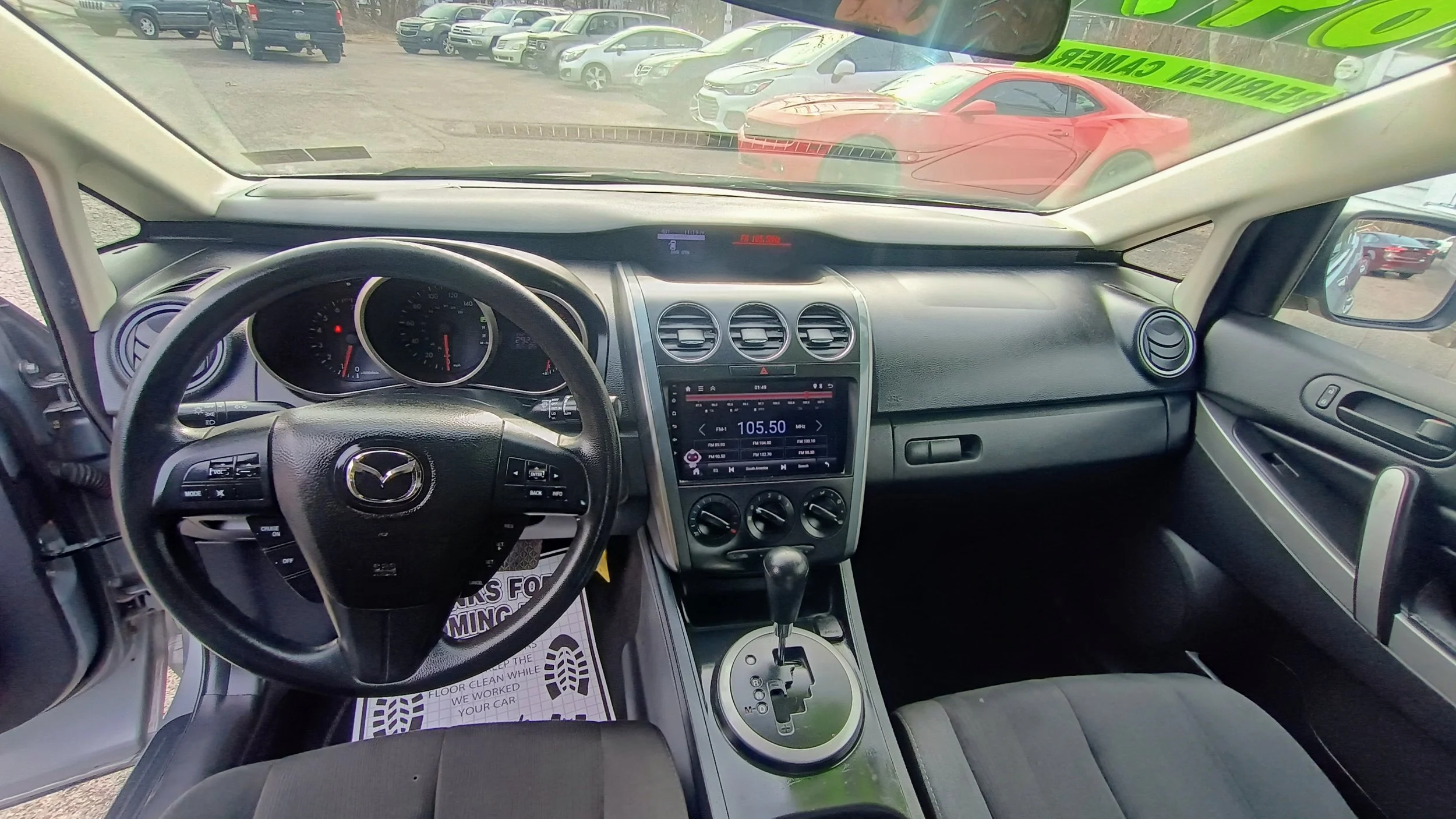 Interior of a Mazda vehicle showing the dashboard, steering wheel, center console with radio tuned to 105.50 FM, and gear shift, with a parking lot and red sports car visible through the windshield.